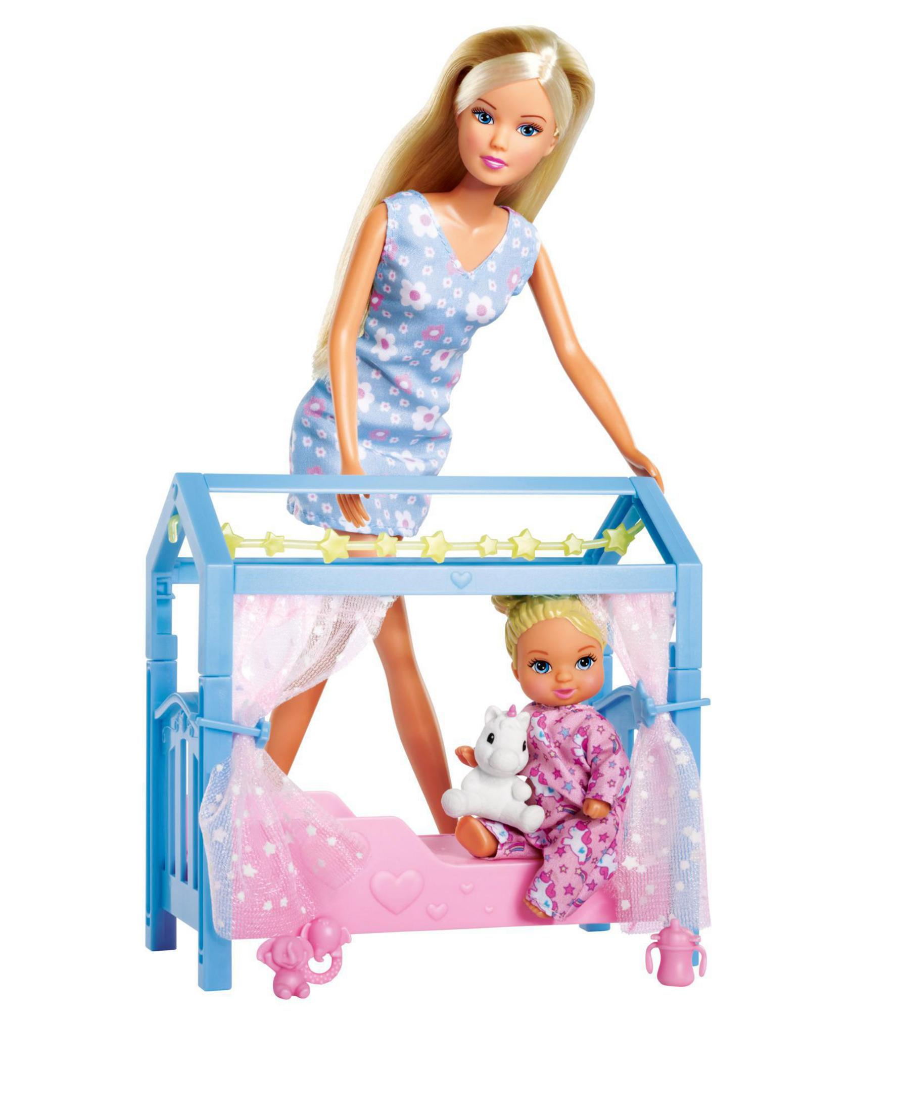 Blonde Puppe steht, mit einer kleineren Puppe in einem blauen Bett. Die Szene ist vor weißem Hintergrund.