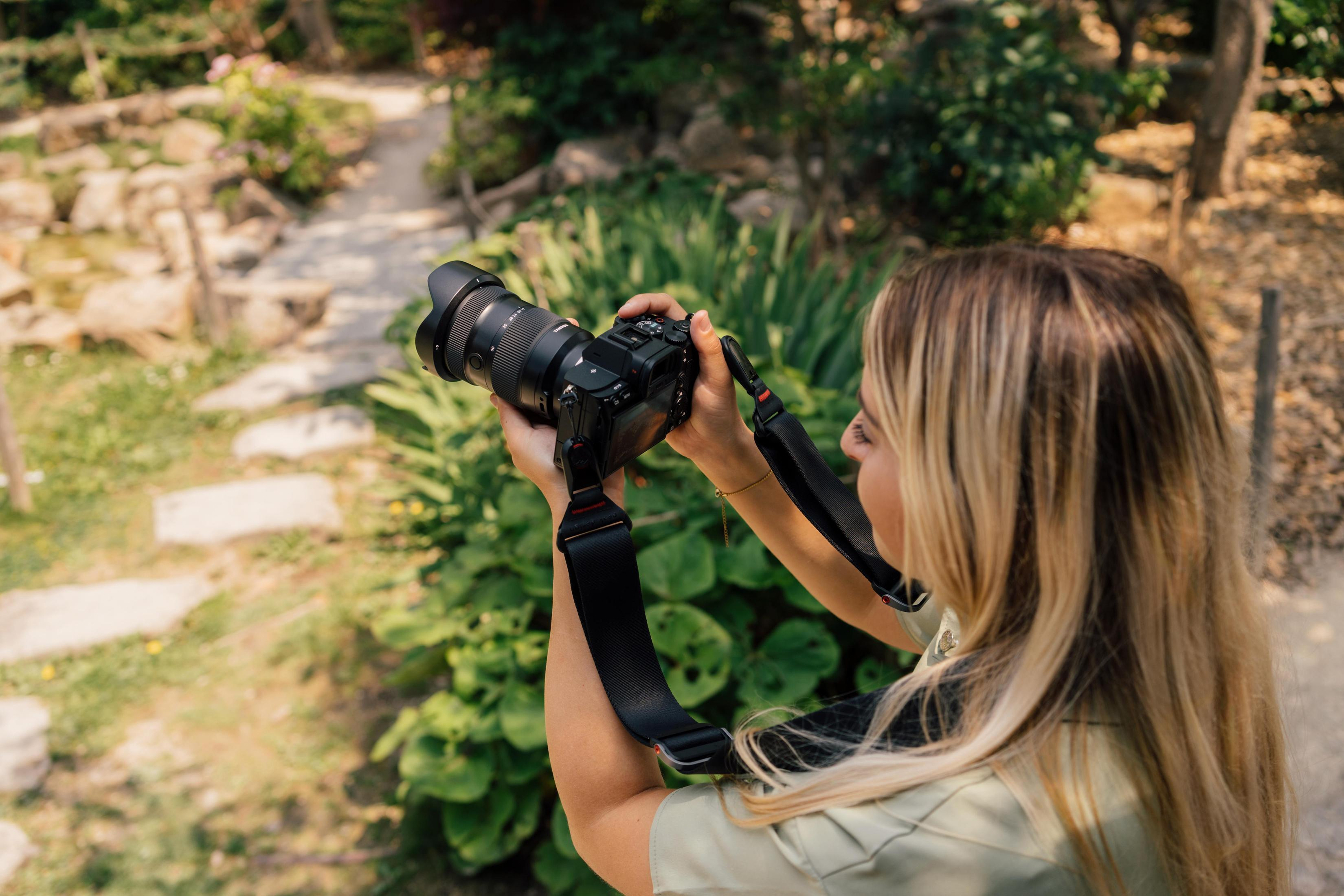 Blondynka fotografuje scenę w ogrodzie aparatem. Zielone rośliny i ścieżka w tle.