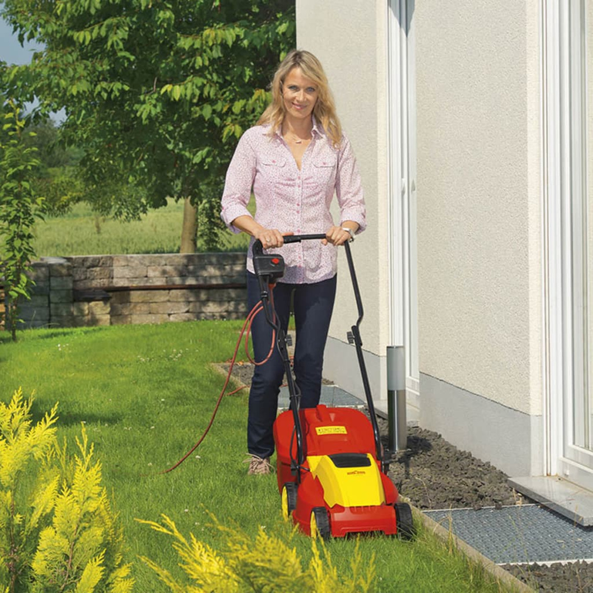 Eine Frau, die einen rot-gelben Rasenmäher auf einem grünen Rasen in der Nähe eines Hauses benutzt.
