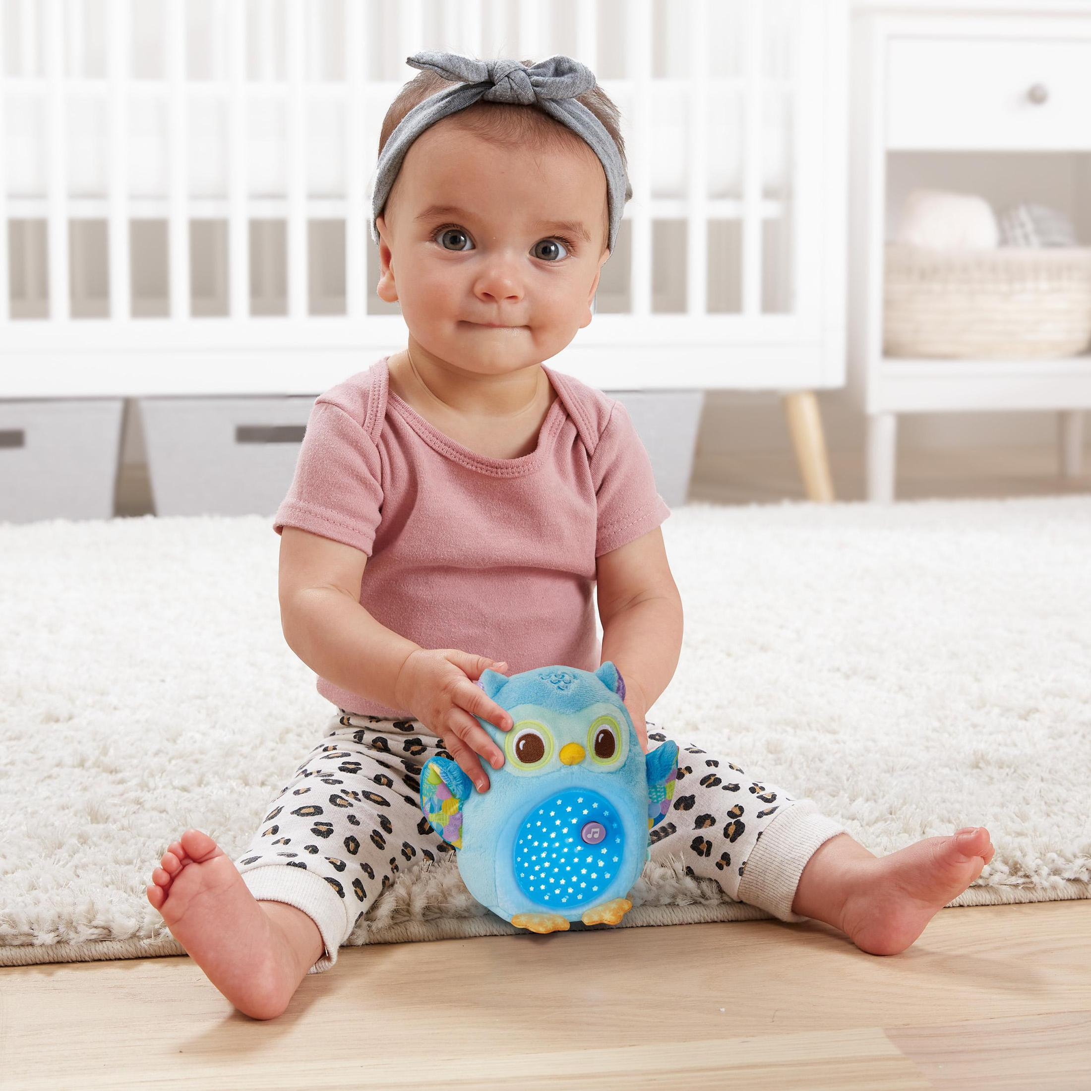 Ein Baby sitzt auf einem Teppich und hält ein blaues Eulen-Spielzeug. Das Baby lächelt und trägt ein Stirnband.