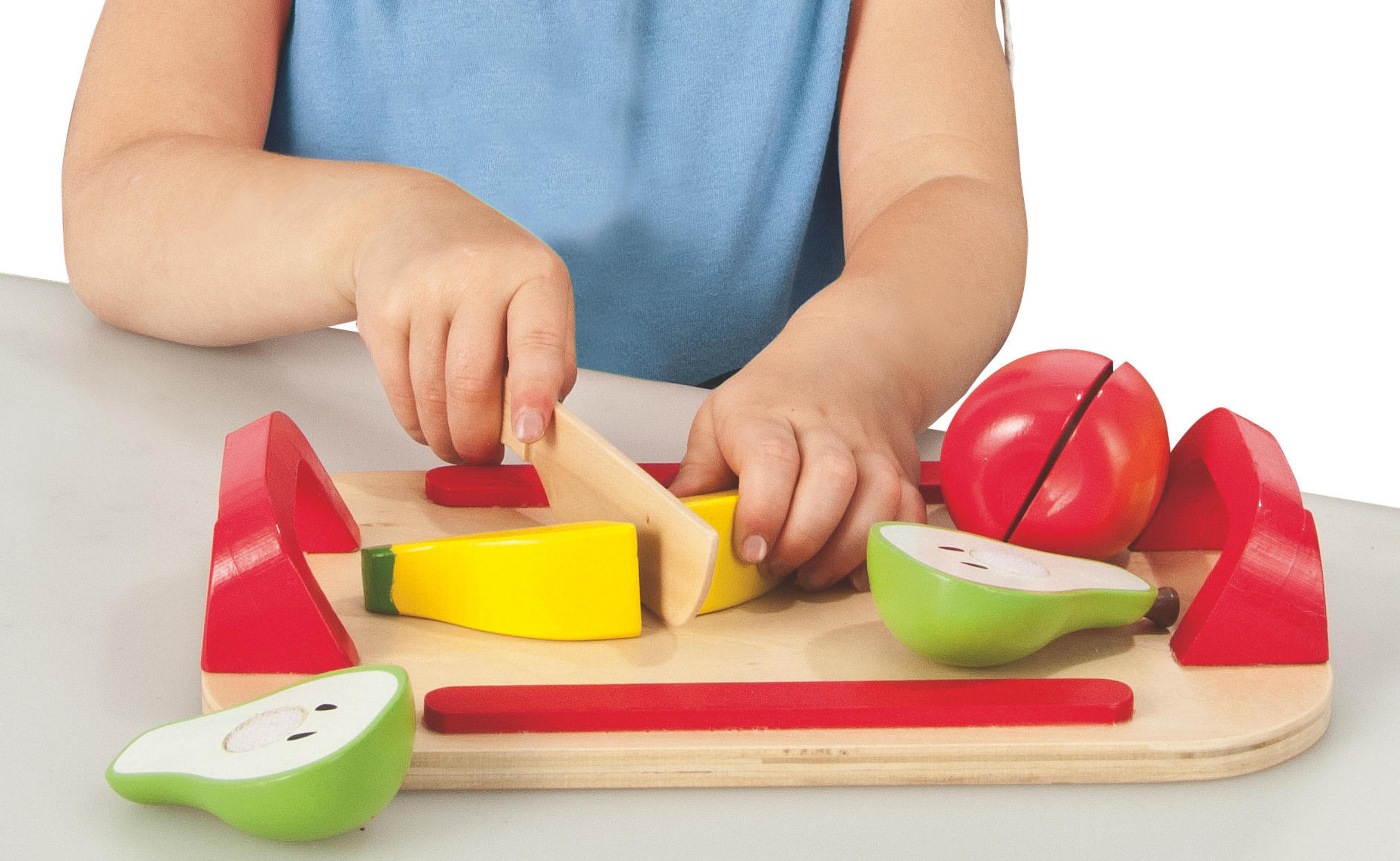 Kind schneidet Spielzeugobst mit einem Holzmesser auf einem Holzschneidebrett. Rotes und gelbes Obst.