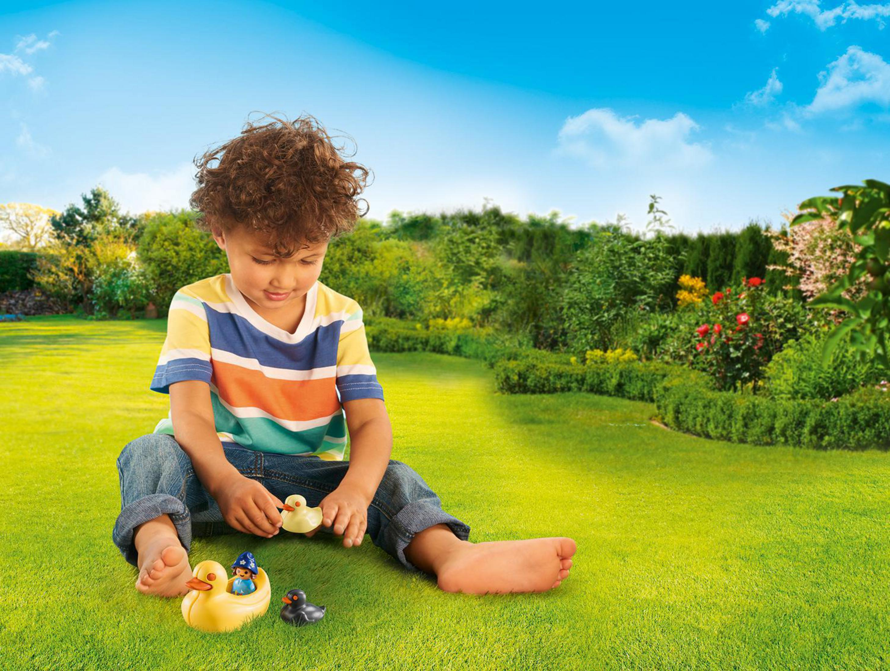 Ein Junge spielt mit Spielzeugenten auf grünem Gras, blauer Himmel im Hintergrund.