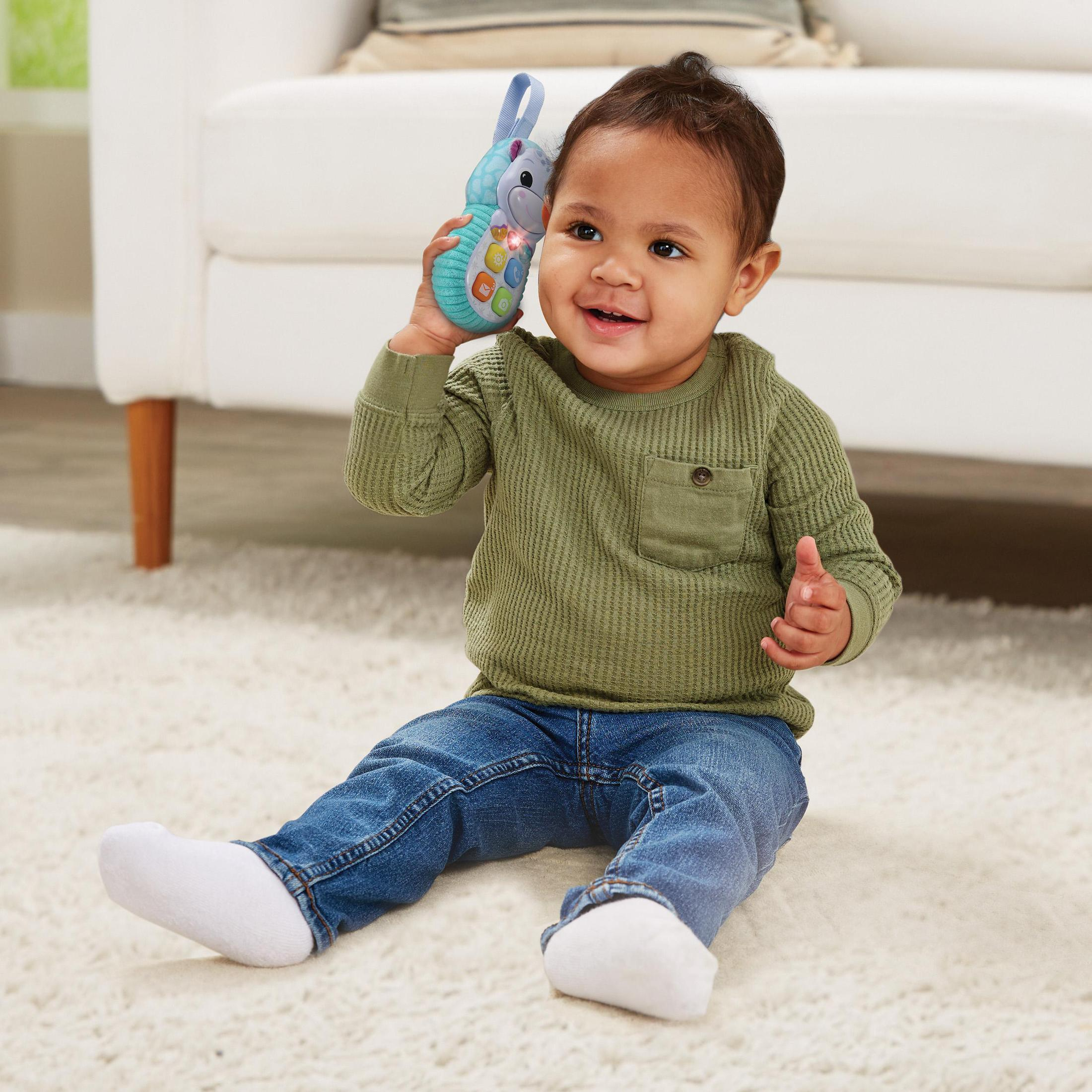 Ein Baby lächelt und hält ein Spielzeugtelefon. Es sitzt auf einem weißen Teppich, trägt ein grünes Hemd und blaue Jeans.