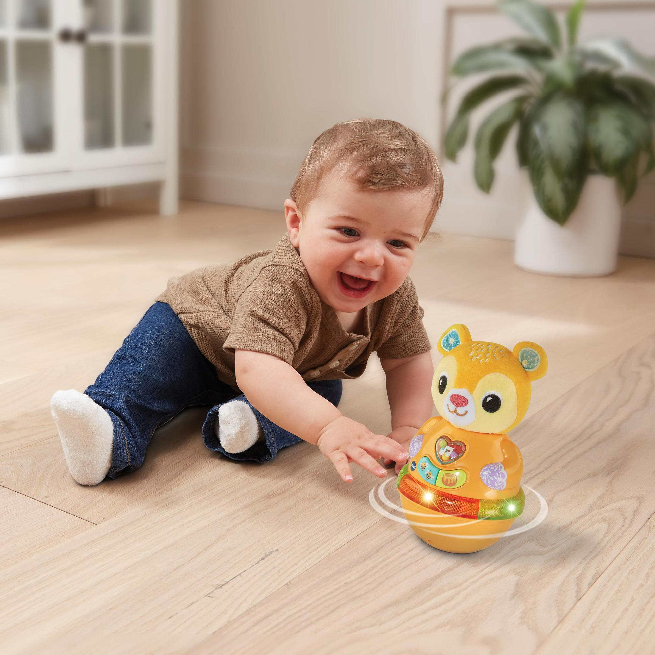 Lachendes Baby krabbelt auf einen Spielzeugbären zu. Das Spielzeug ist orange, mit Lichtern, Baby in braunem Hemd.