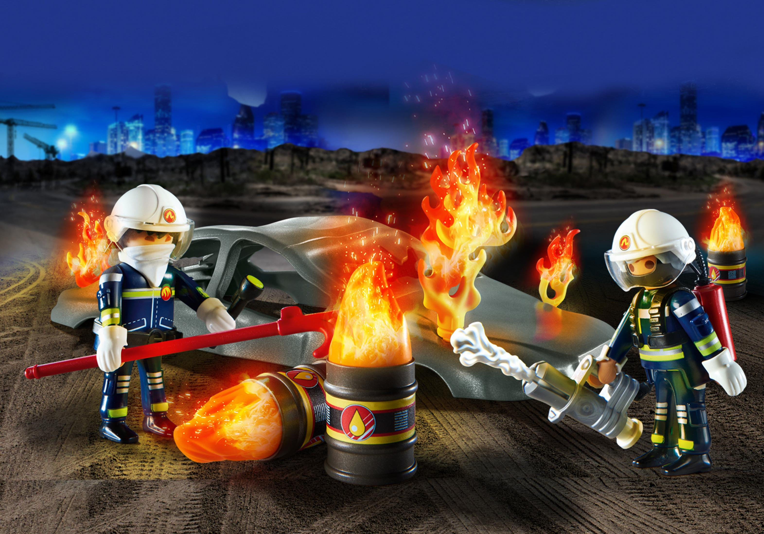 Bomberos Playmobil con fuego y un coche en llamas por la noche. Skyline al fondo.