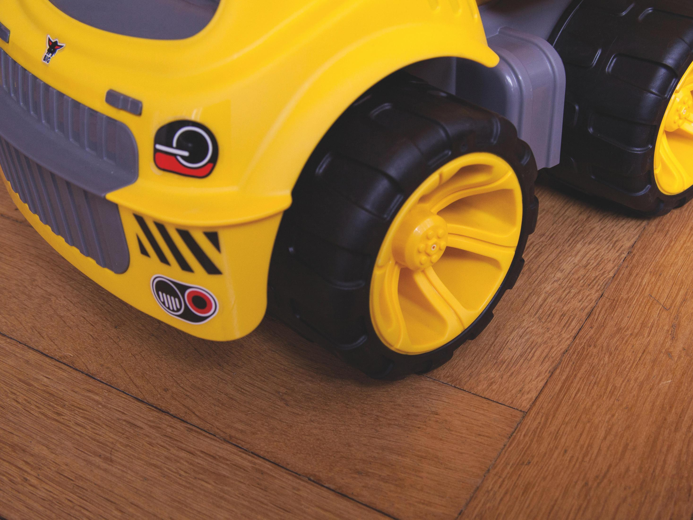 Żółte i czarne koło zabawkowego samochodu ciężarowego. Czarna opona, żółta felga i czarno-żółta naklejka. Drewniana podłoga.