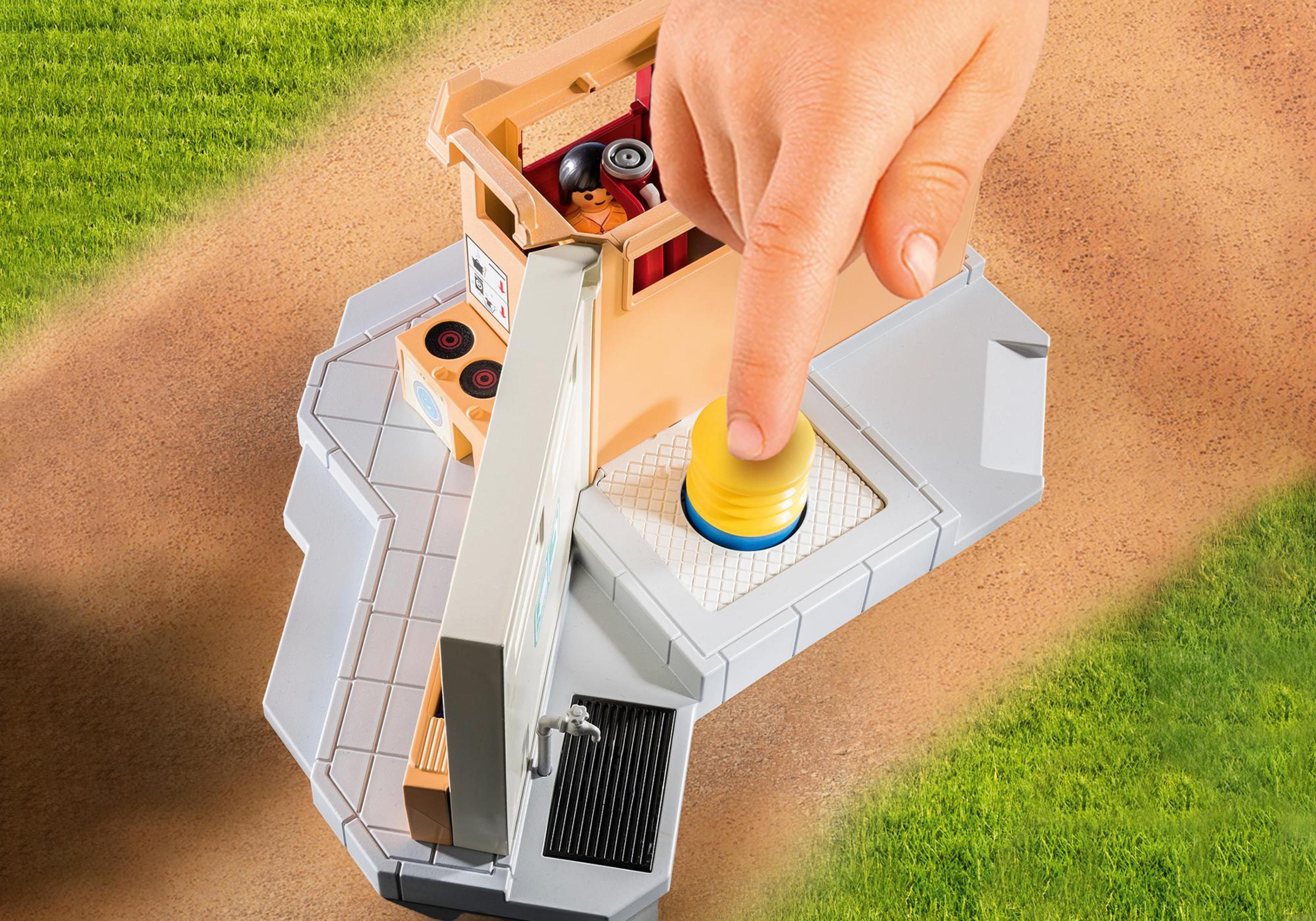Una mano che preme un pulsante giallo su un set di giocattoli Playmobil, con un piccolo edificio.