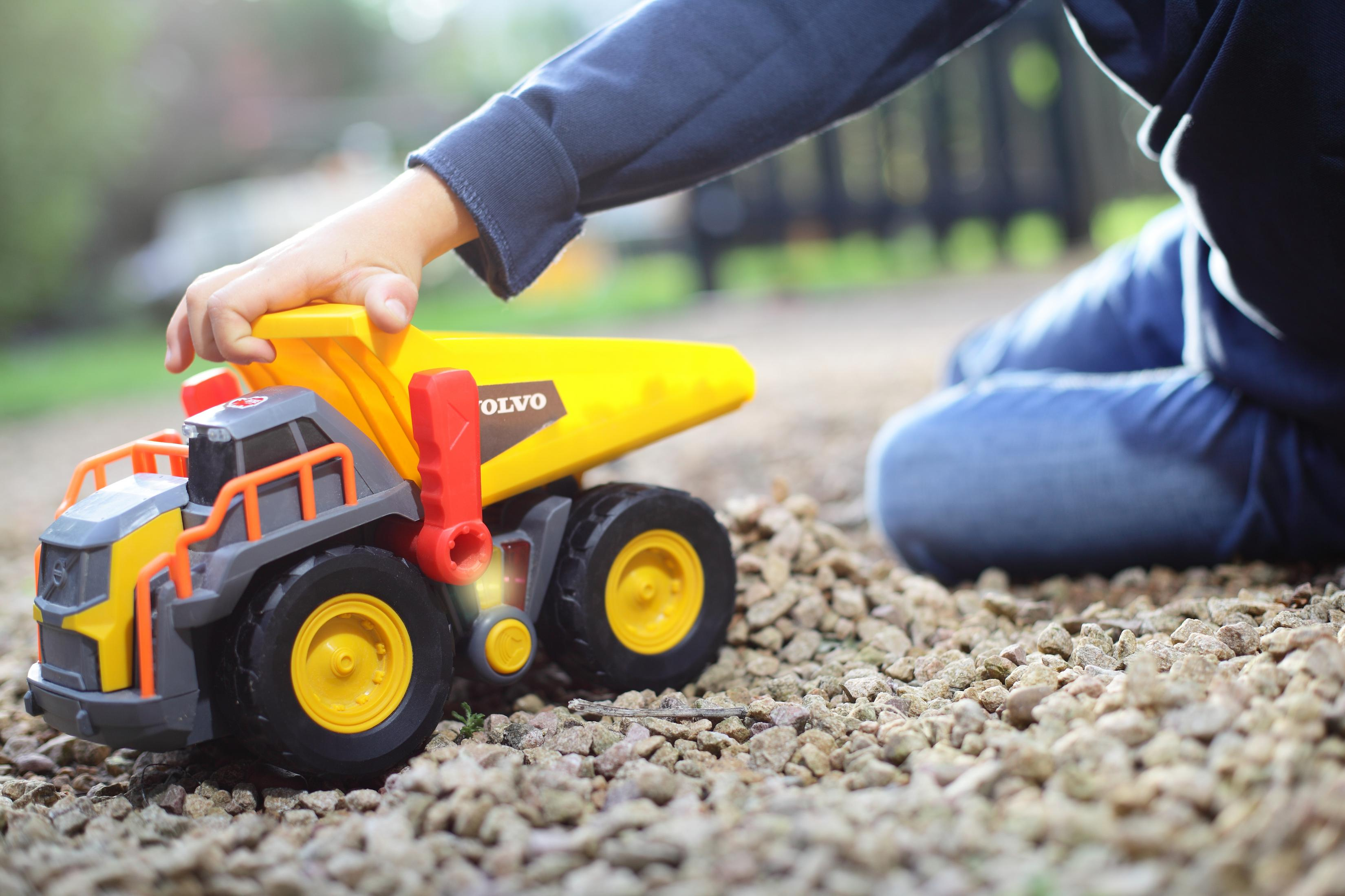 Ręka dziecka bawi się żółto-szarą zabawką ciężarówką Volvo na żwirze.