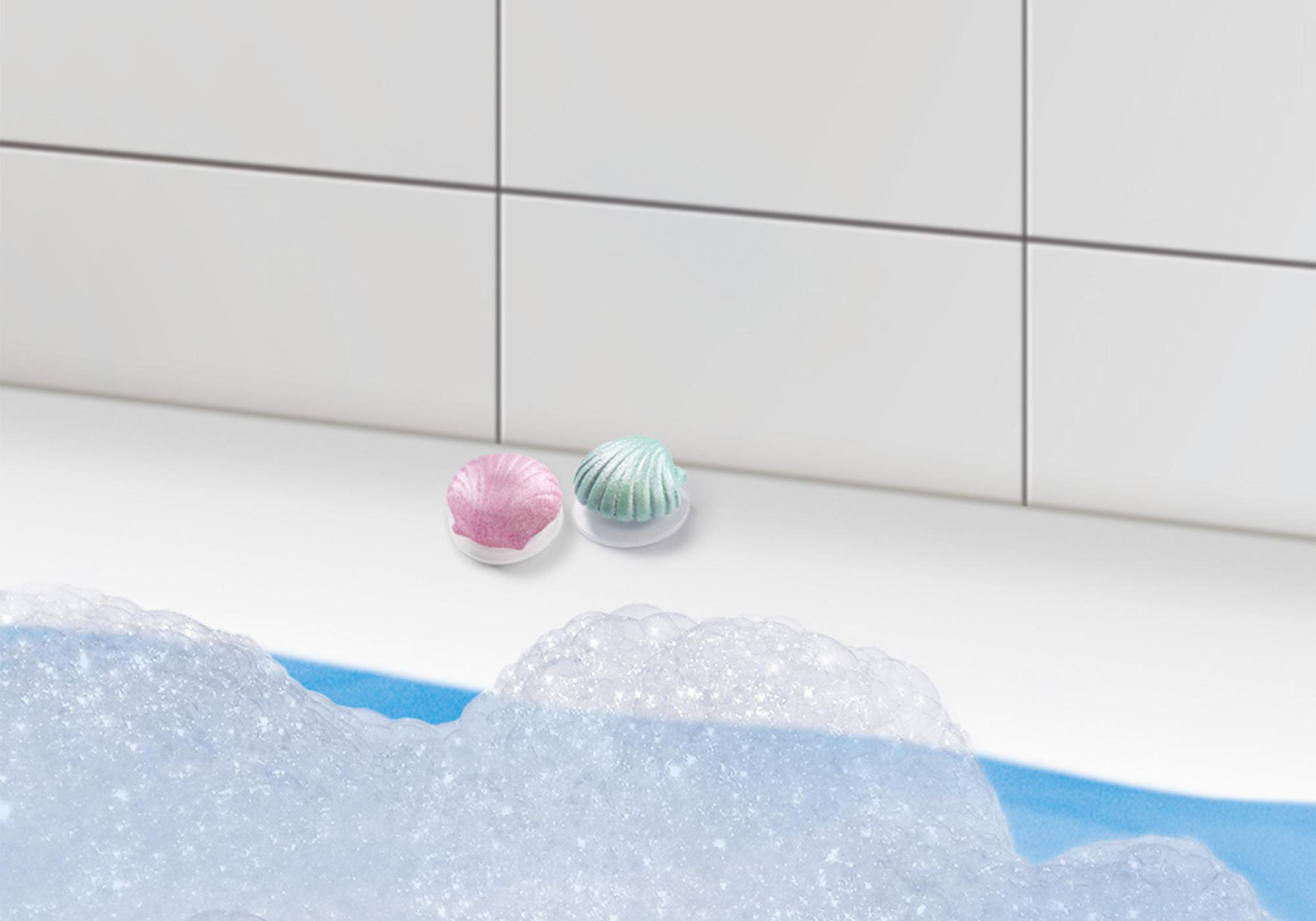 Zwei muschelförmige Badebomben, rosa und grün, in der Nähe einer weißen Fliesenwand und schaumigem Wasser.
