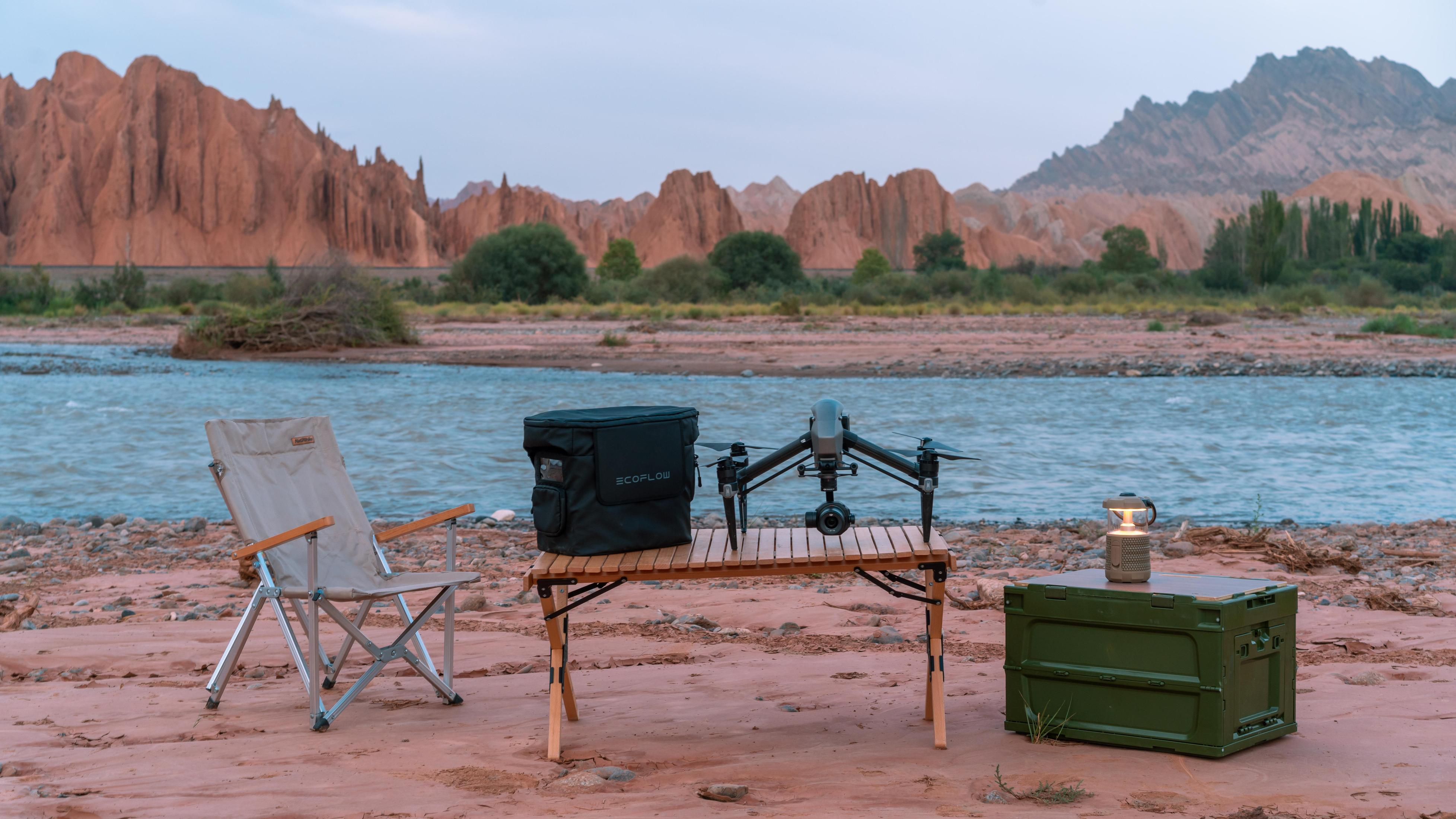 Dron, krzesło kempingowe, stół i pojemnik stoją nad rzeką. W tle góry.
