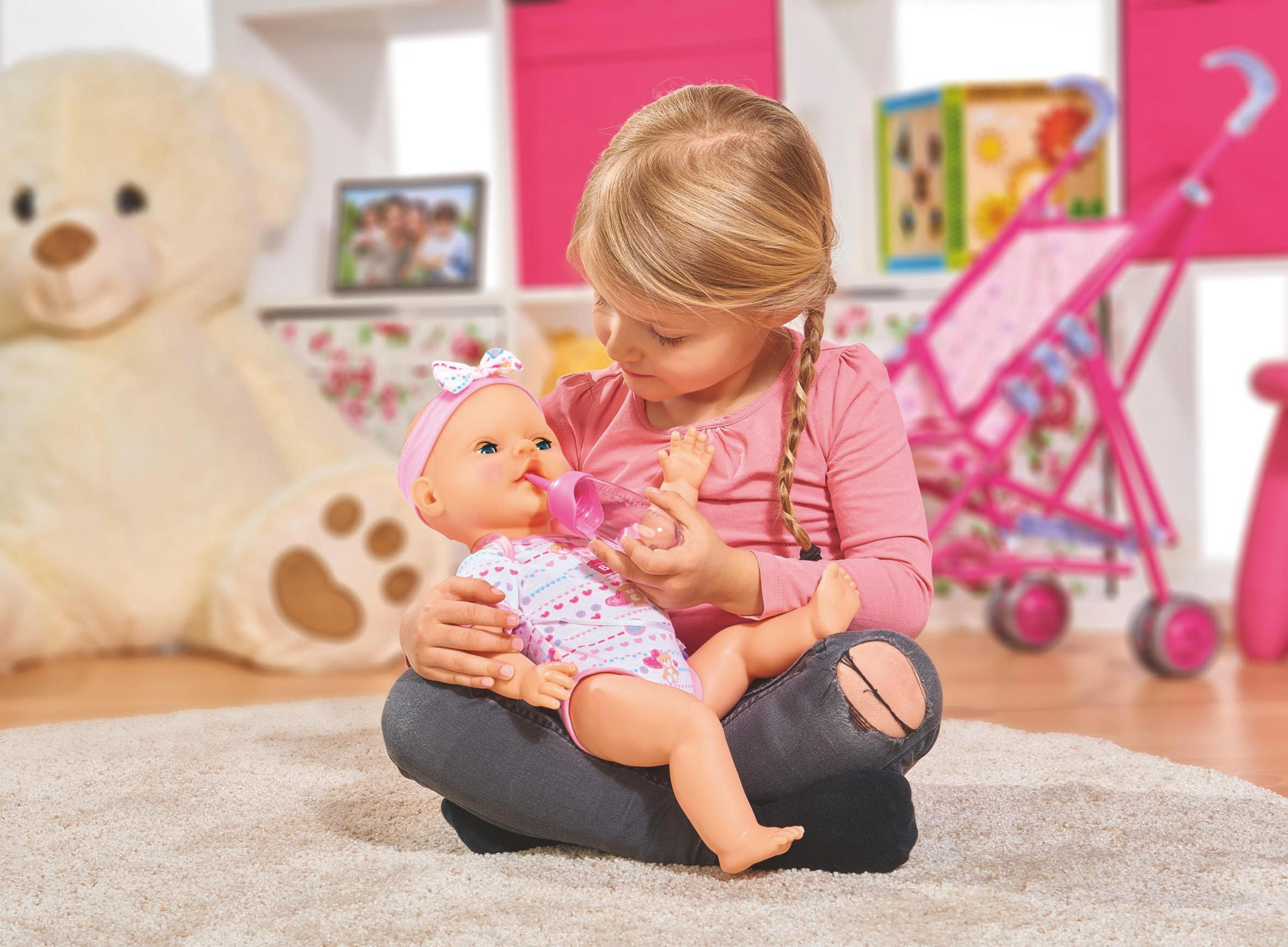 Ein Mädchen füttert eine Babypuppe mit einer Flasche, neben einem rosa Kinderwagen.