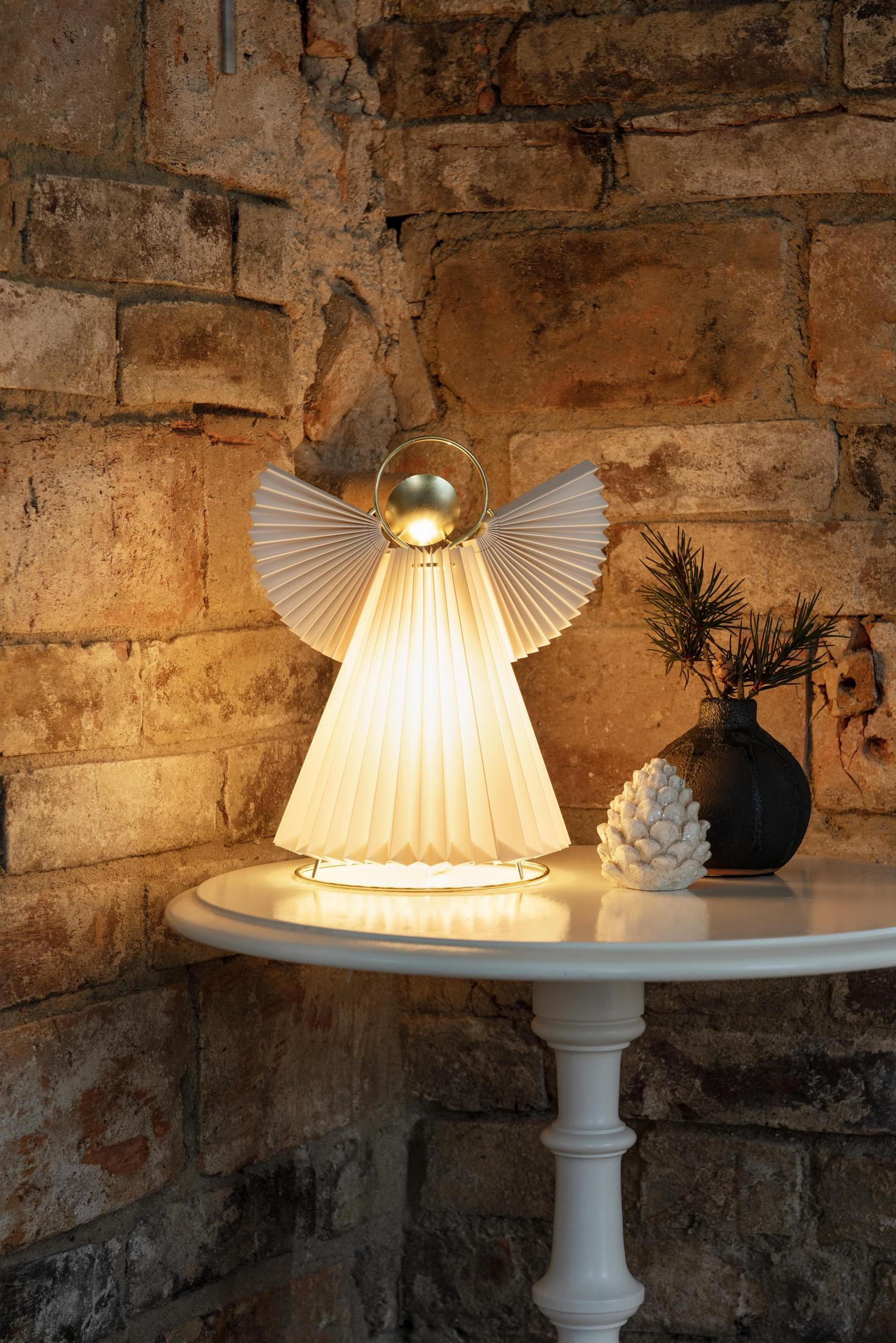 Eine engelartige Lampe auf einem weißen Tisch, mit Backsteinmauer im Hintergrund. Sie hat einen goldenen Heiligenschein.