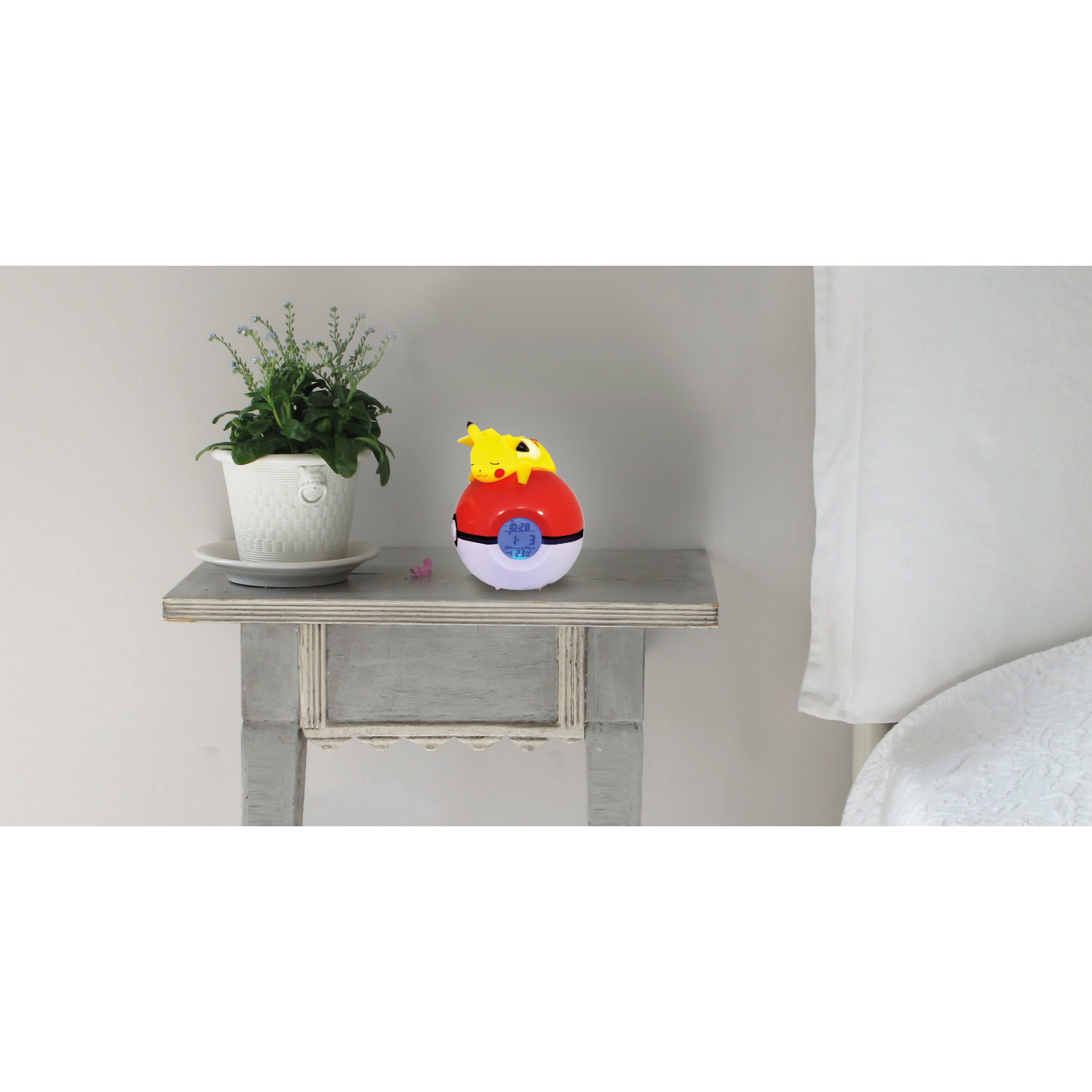 Budzik Pikachu na stole, z rośliną i łóżkiem w tle.