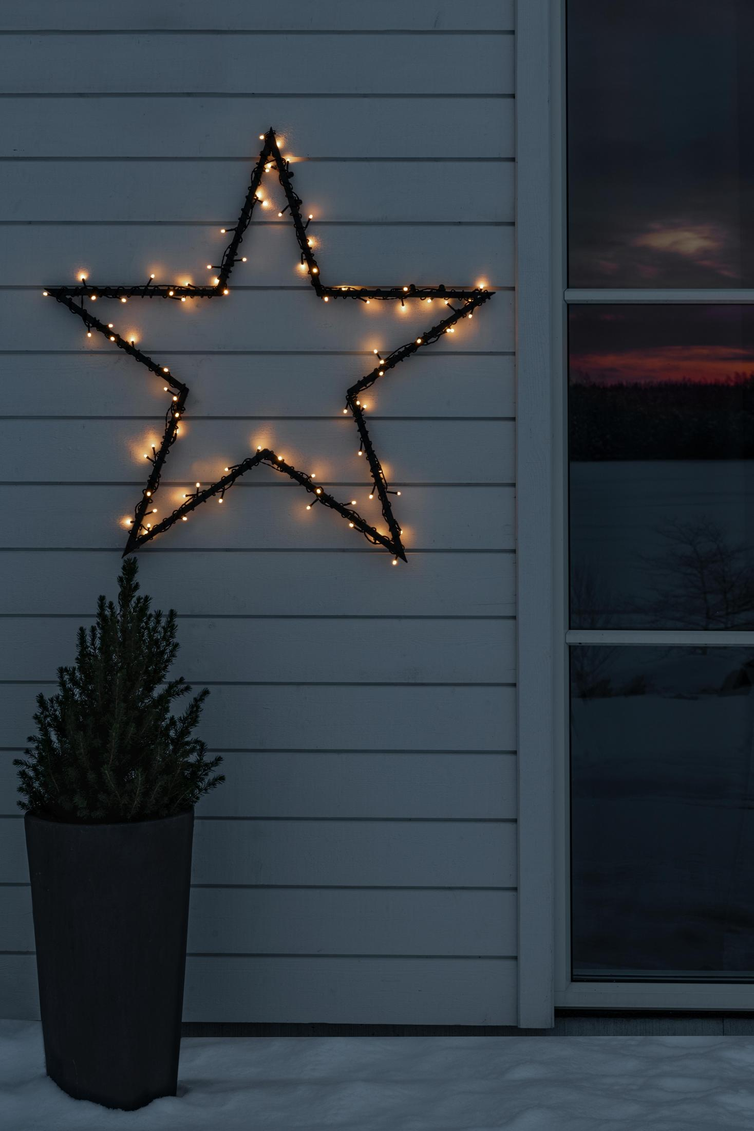 Ein sternförmiger Weihnachtslicht-Display an einem weißen Haus, mit einem Topfbaum davor.