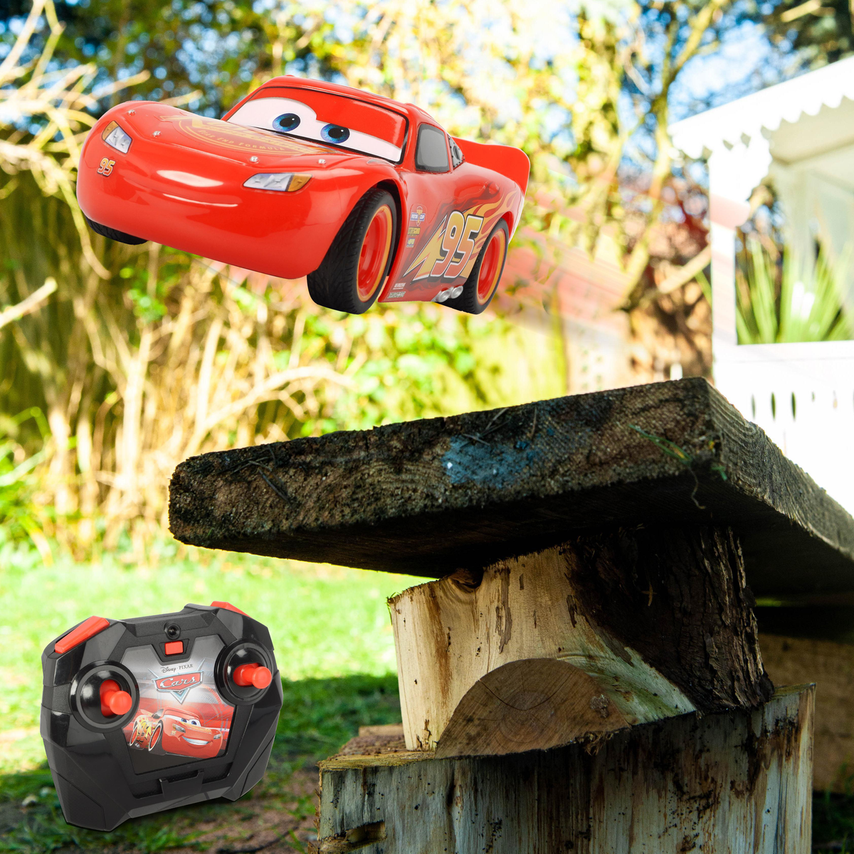 Coche Lightning McQueen saltando sobre un banco de madera, controlado por un mando. Fondo: árboles verdes y una casa.