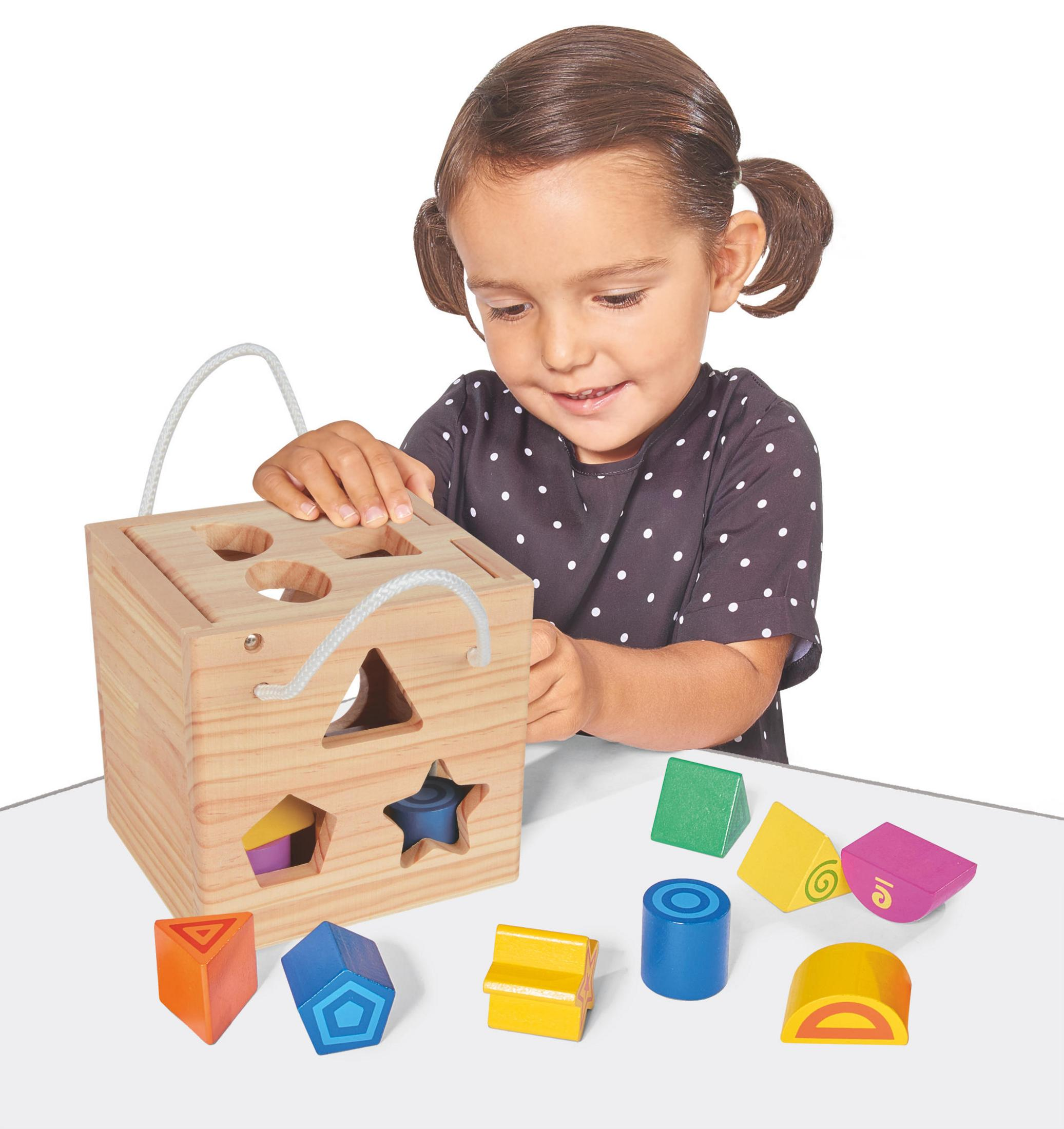 Ein Mädchen spielt mit einem hölzernen Formensortierspielzeug. Bunte Formen und eine Holzkiste sind sichtbar.