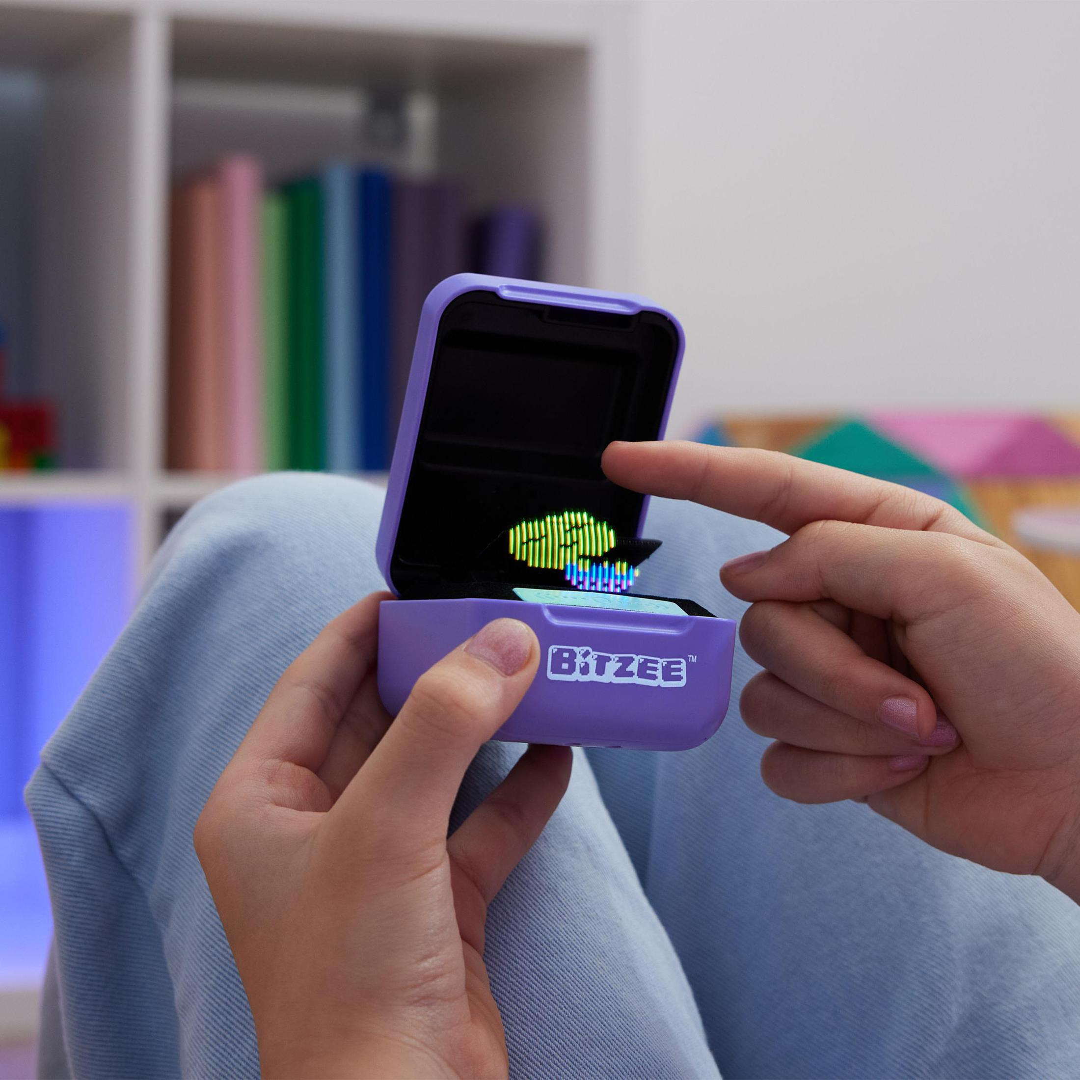 Hände öffnen ein lila Bitzee-Spielzeug, das ein grün-blaues Bild zeigt. Hintergrund: ein Regal mit Büchern.
