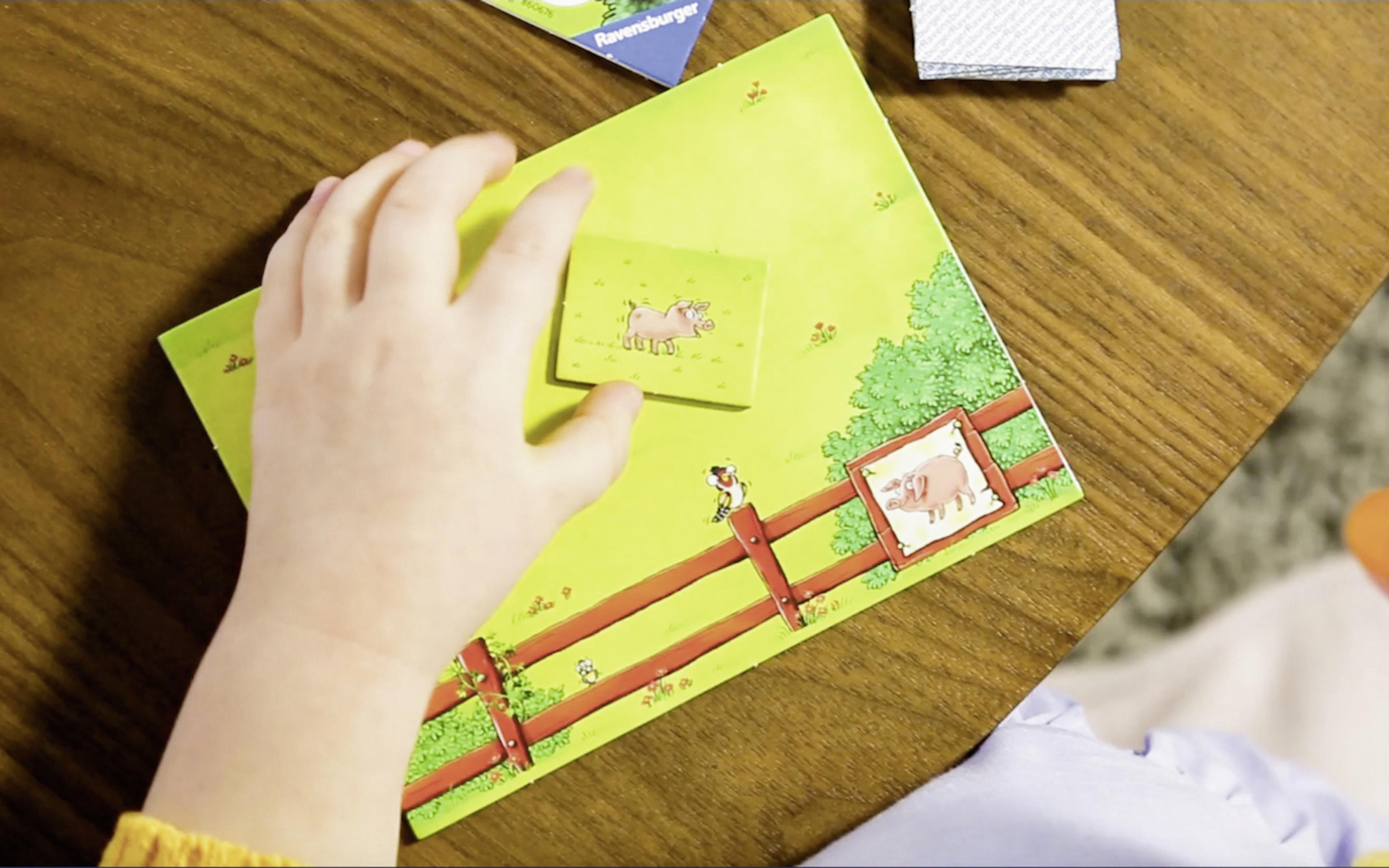 Die Hand eines Kindes platziert ein Schweinepuzzle-Teil auf einer Bauernhofszene. Andere Teile sind auf dem Tisch.