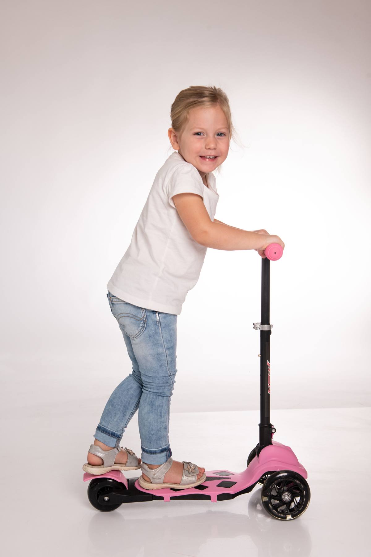Ein Mädchen auf einem rosa Roller lächelt, stehend auf einem weißen Hintergrund. Sie trägt Sandalen und Jeans.