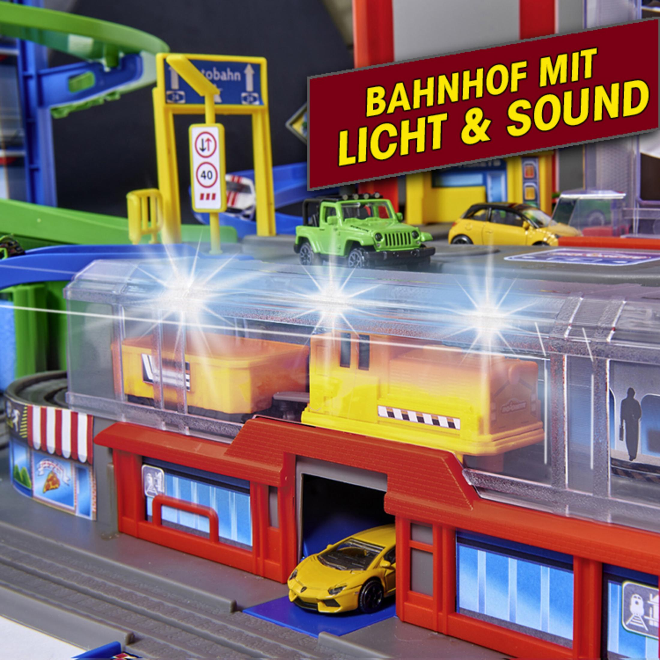 Ein Modelleisenbahn-Set mit einem gelben Zug, einem grünen Jeep und einem gelben Auto. Schild sagt 'BAHNHOF'.