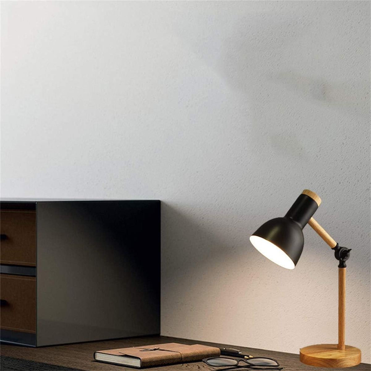 Schwarze Schreibtischlampe auf Holzständer. Notizbuch, Brille und schwarzer Schrank im Hintergrund.