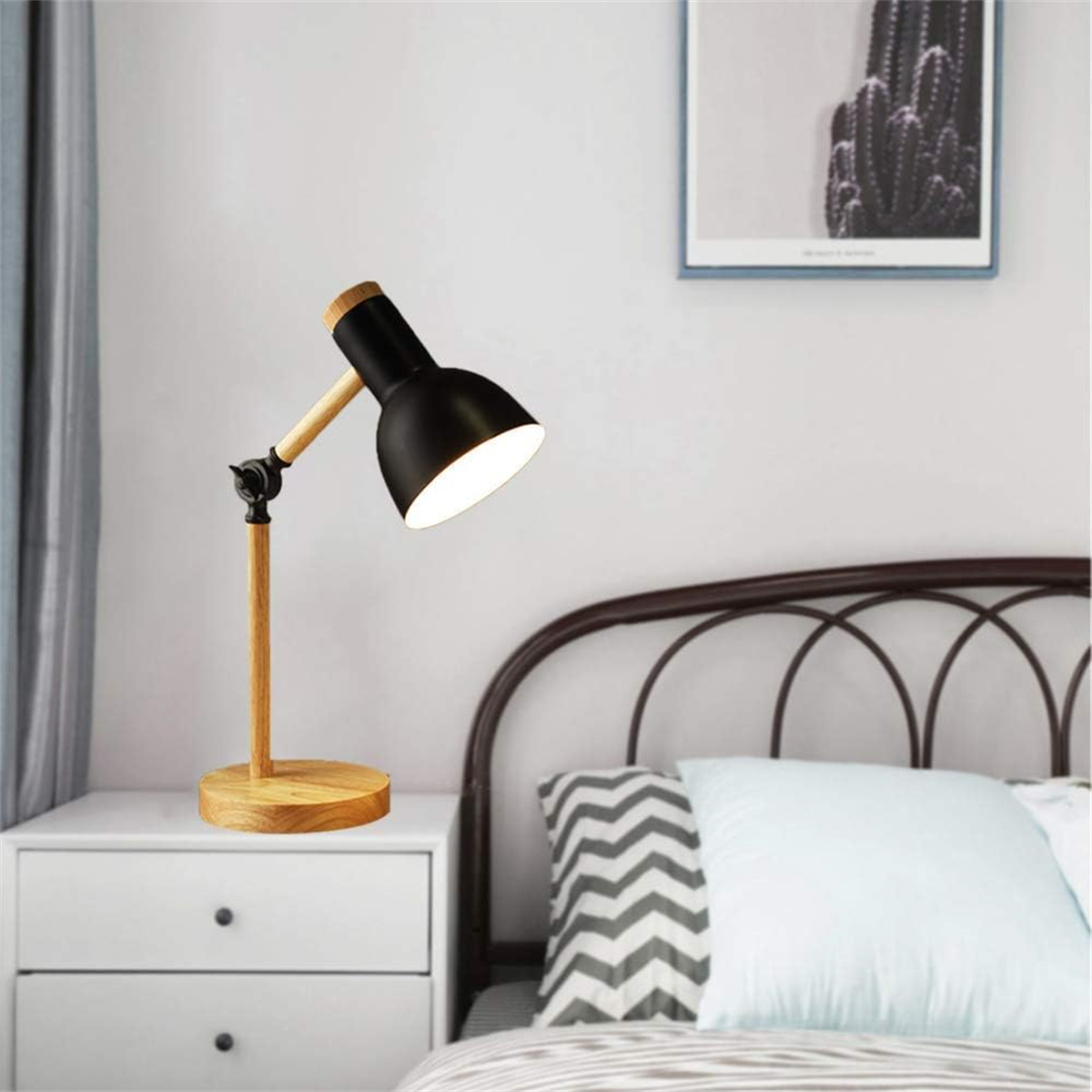 Eine schwarze Schreibtischlampe mit Holzfuß, auf ein Bett mit Nachttisch gerichtet.