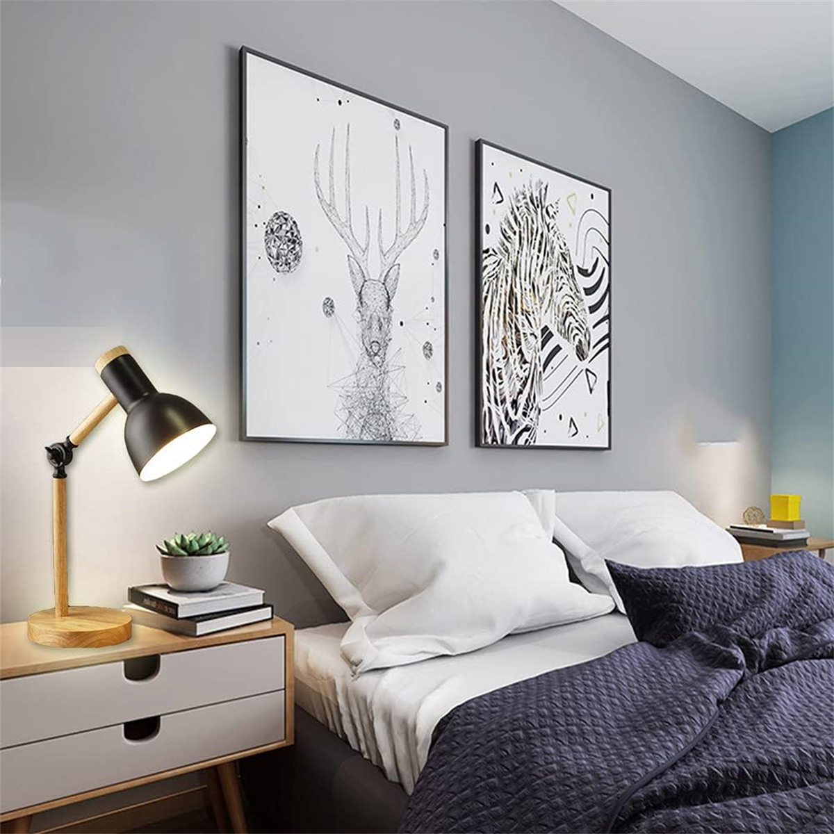 Eine schwarze und hölzerne Schreibtischlampe steht auf einem Nachttisch mit Kunstdrucken und Büchern. Ein Bett ist im Hintergrund.