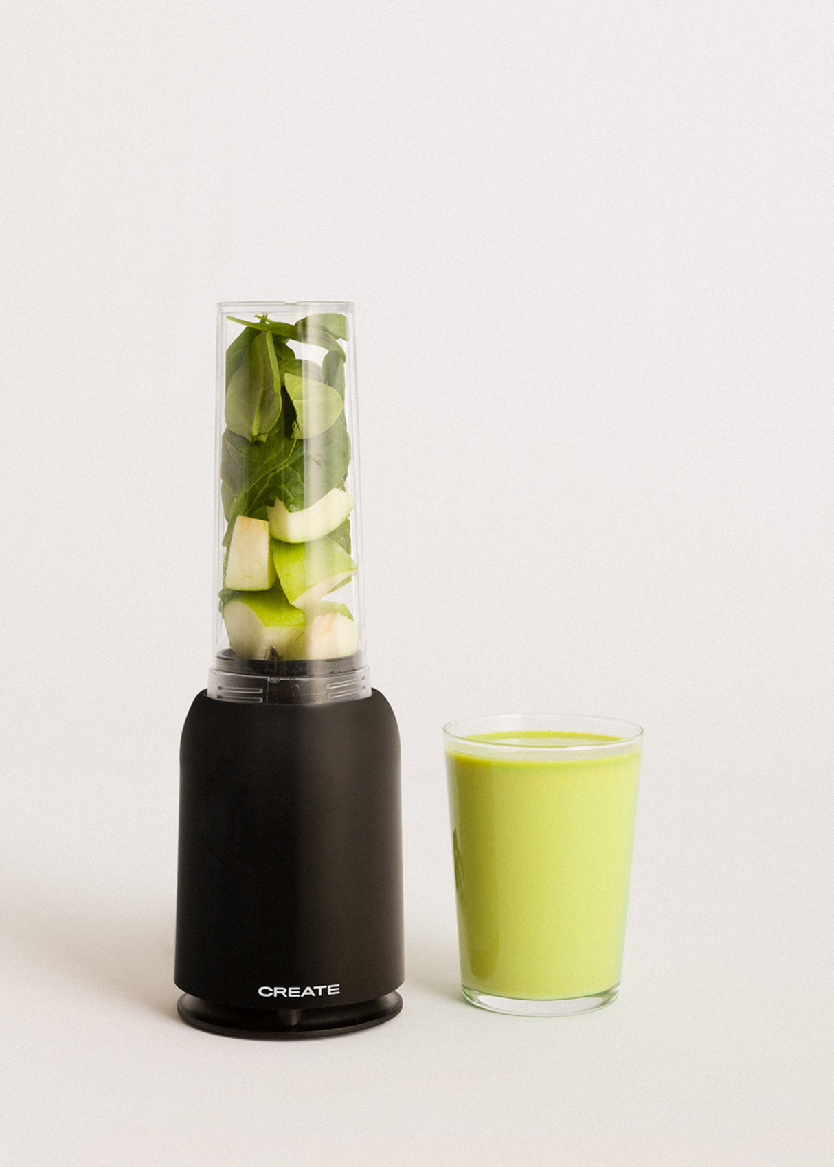 Una licuadora negra con frutas y verduras dentro. Un vaso de batido verde junto a ella sobre blanco.