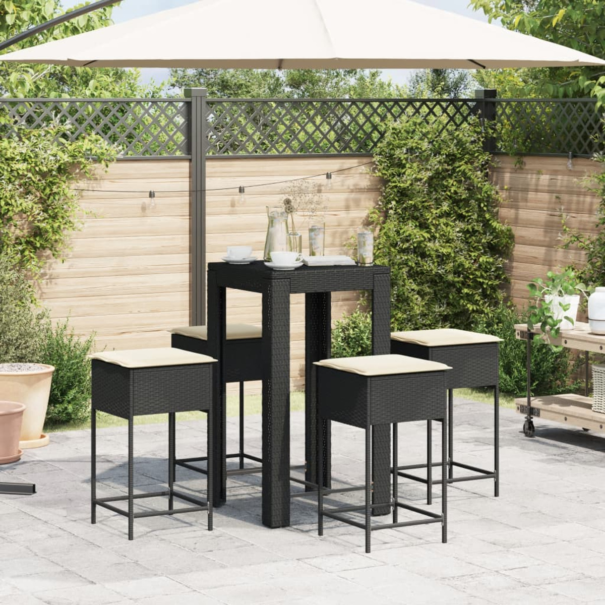 Set tavolo e sgabelli in vimini nero da esterno con cuscini beige, sotto un ombrello beige, su un patio.