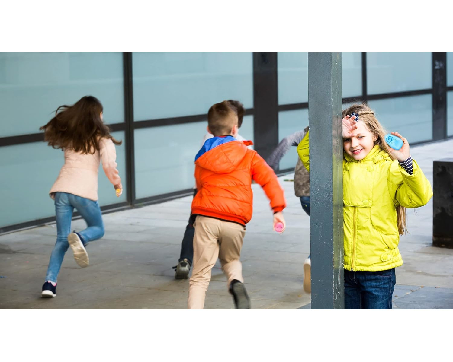 Kinder spielen, ein Mädchen in gelber Jacke lehnt an einer Stange, lächelnd.