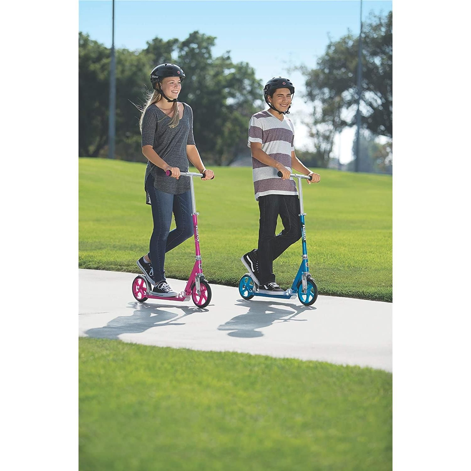 Deux personnes font du scooter sur un sentier pavé, avec de l'herbe verte et un ciel bleu.