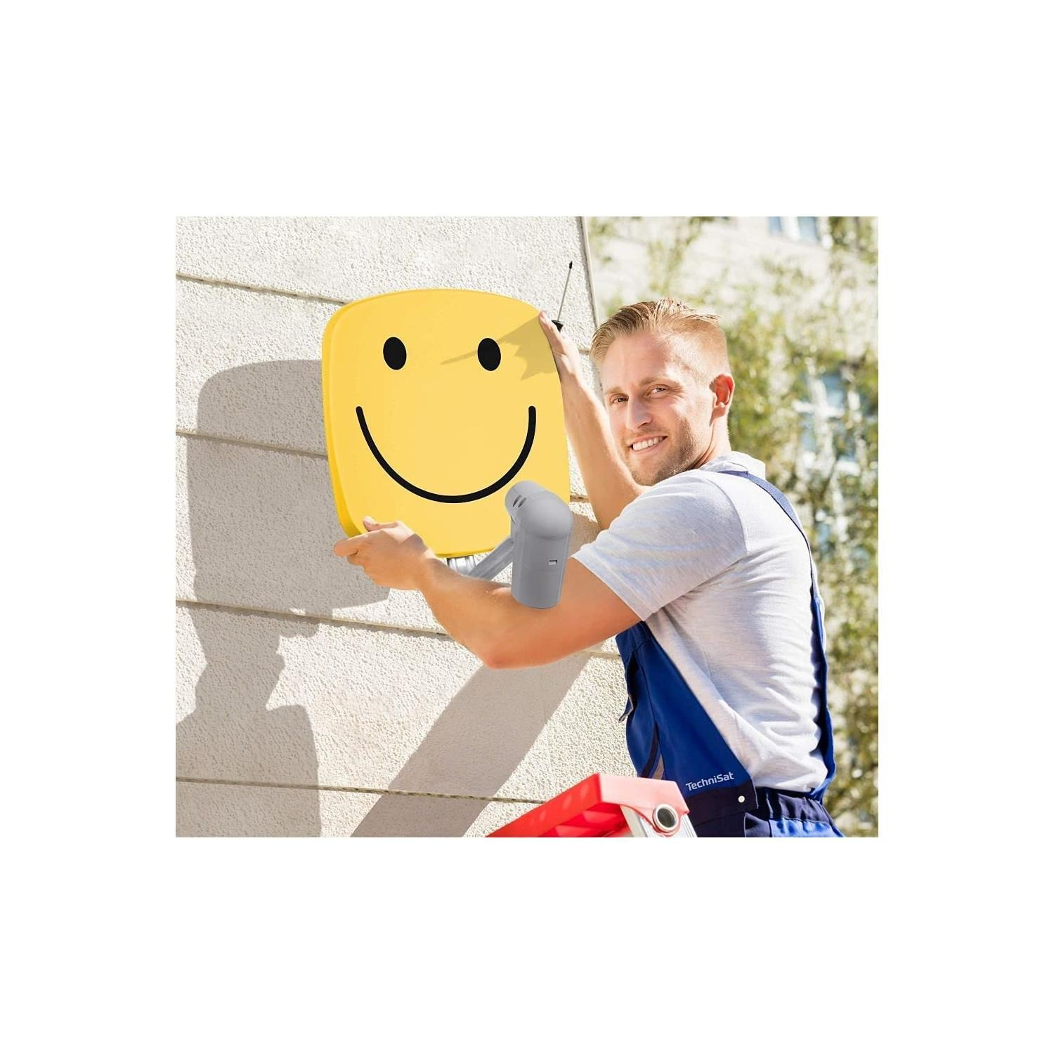 Mann installiert eine gelbe Smiley-Antenne an einer Wand. Blaue Latzhose.