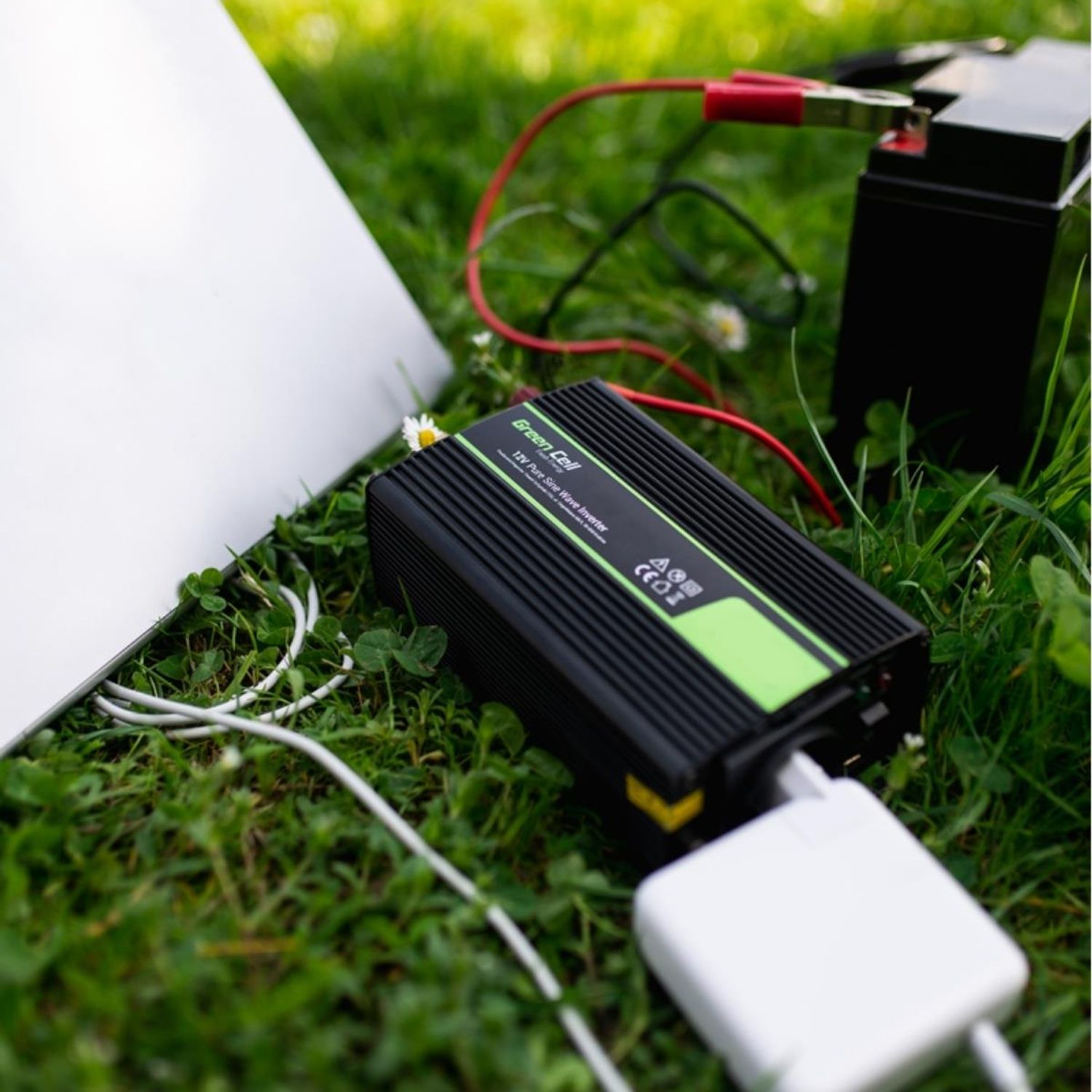Ein schwarzer und grüner Wechselrichter im Gras, an eine Batterie und ein weißes Ladegerät angeschlossen.