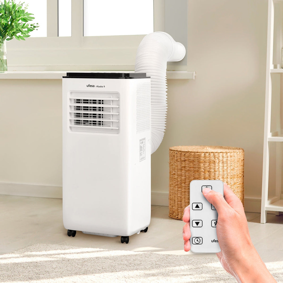 Biały przenośny klimatyzator. Biała rura prowadzi do okna. Pilot w dłoni.