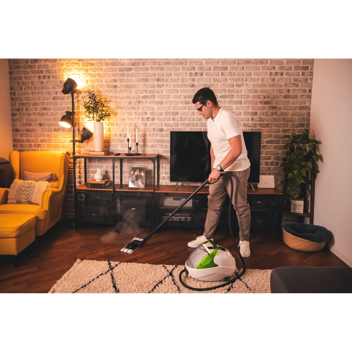 Mężczyzna czyszczący dywan parą za pomocą maszyny. Pokój z telewizorem, kanapą i ceglaną ścianą.