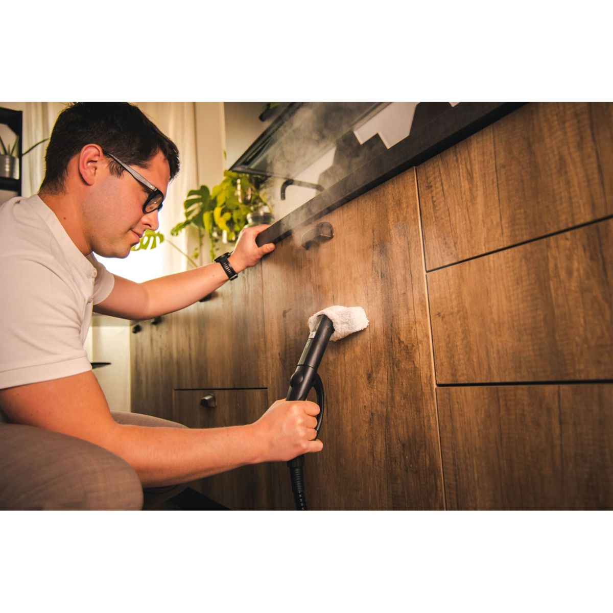 Mężczyzna czyści drewnianą szafkę parą. Ma na sobie okulary i trzyma parownicę.