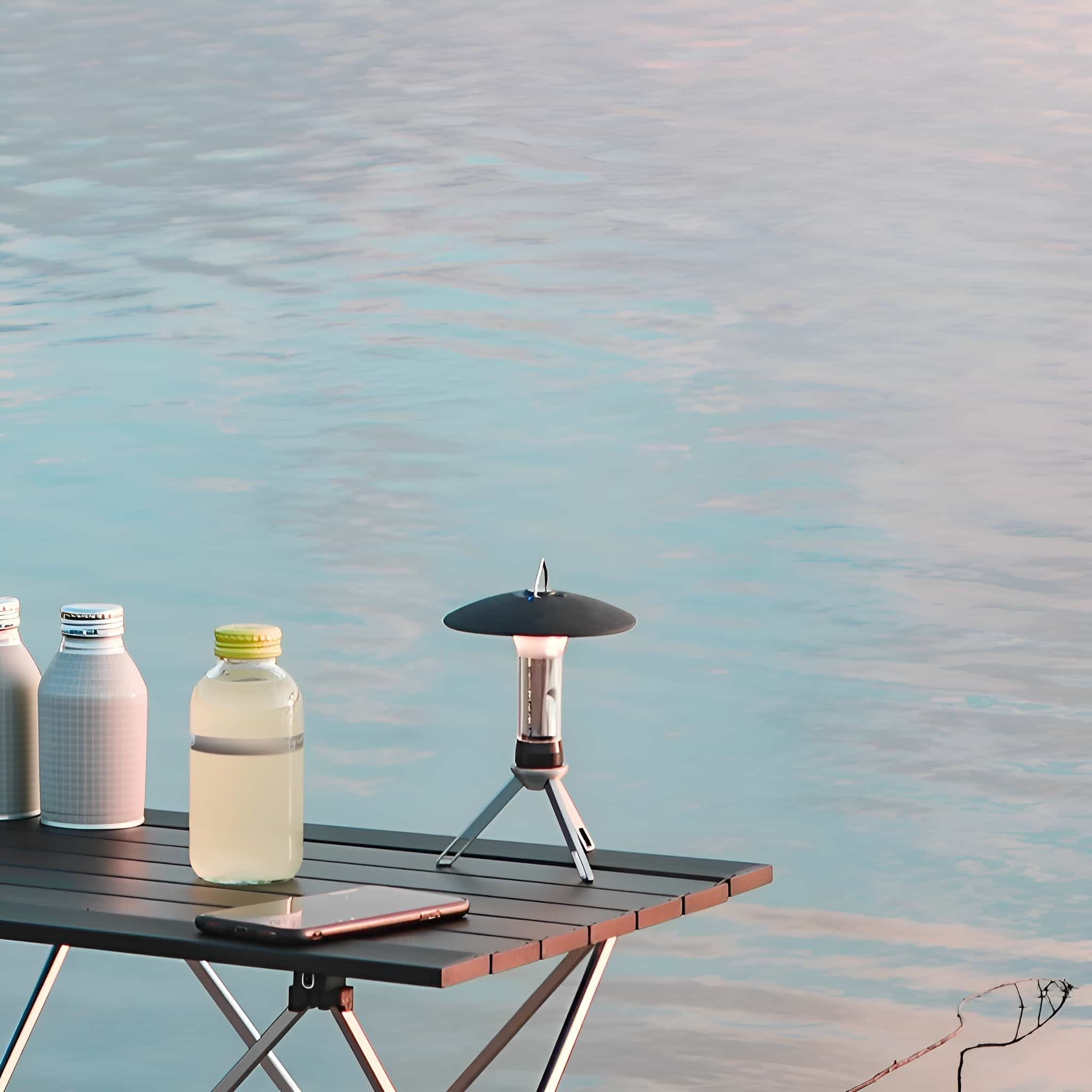 Eine kleine Campinglaterne auf einem Tisch in der Nähe von Wasser, mit Flaschen und einem Smartphone.