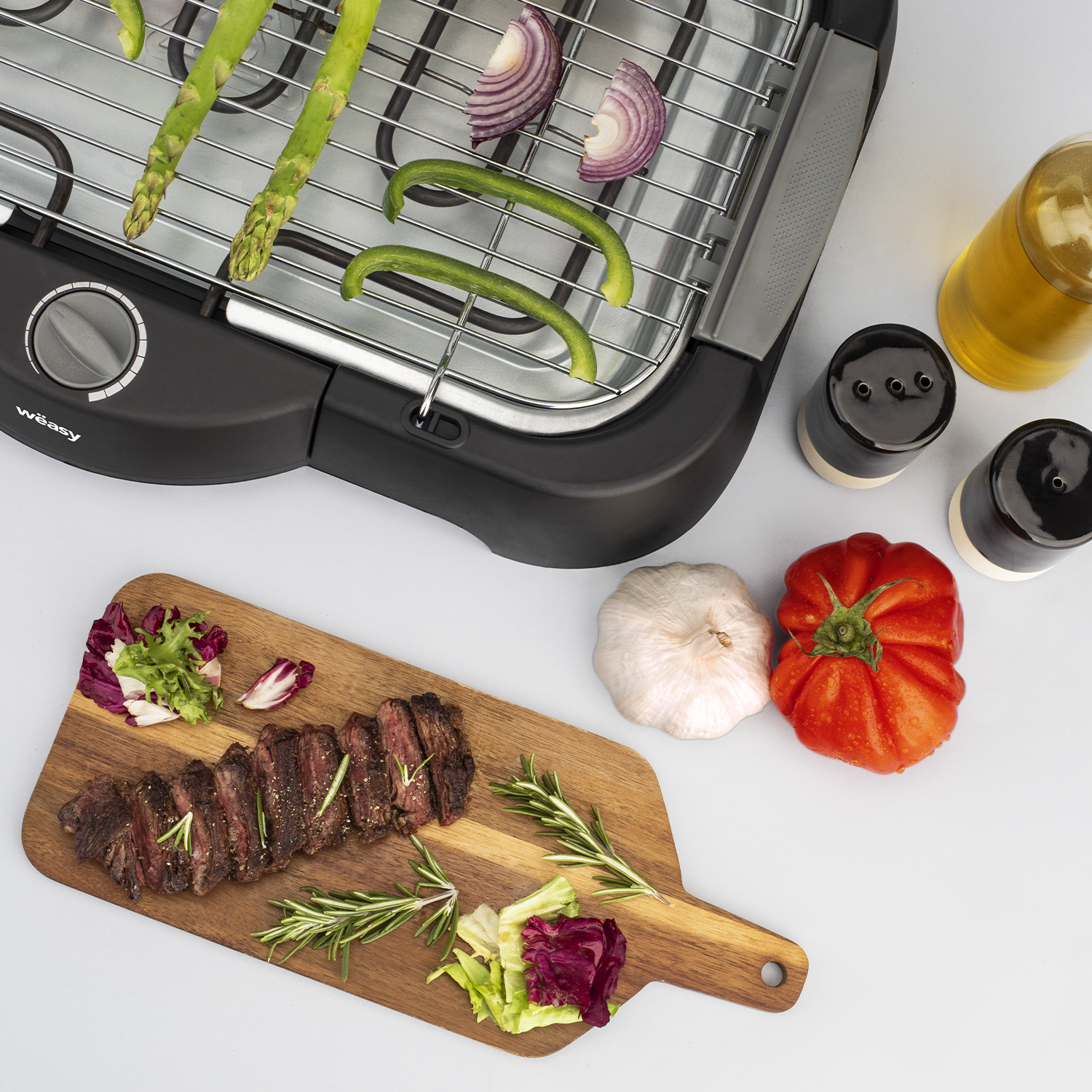 Ein Grill mit Essen: Steak, Spargel, Zwiebeln, Tomaten. Öl- und Salz-/Pfefferstreuer umgeben den Grill.