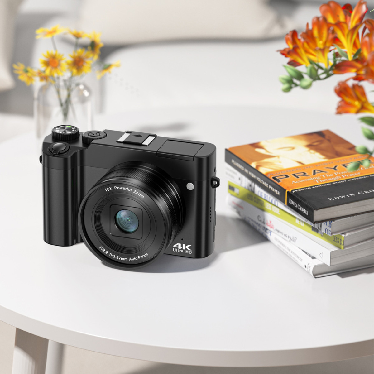 Czarny aparat kompaktowy z obiektywem i przyciskami, obok książek i wazonu z kwiatami. 4K Ultra HD.