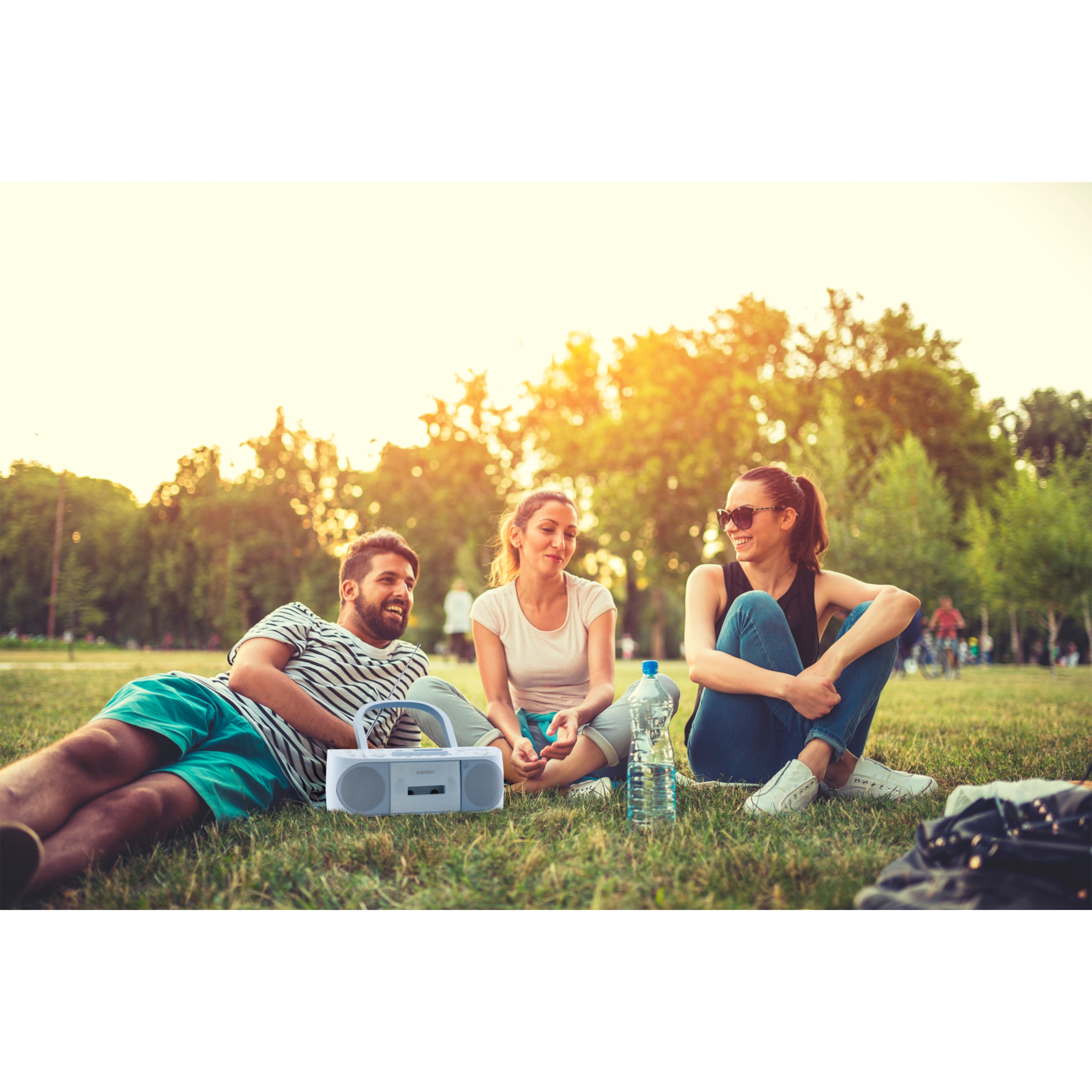 Tre persone sedute sull'erba con una radio. Bottiglia d'acqua visibile. Parco soleggiato.