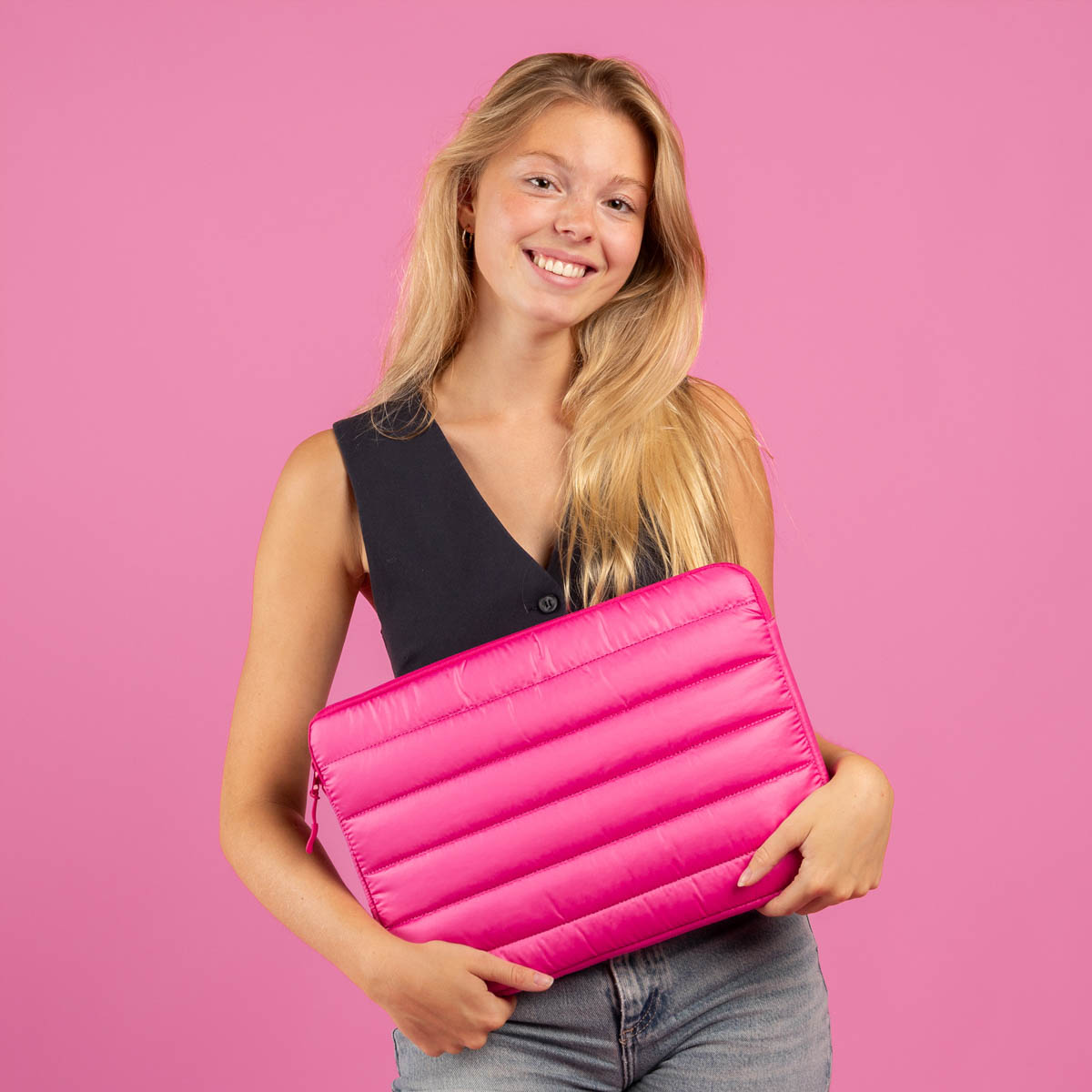 Blonde woman holding a pink padded laptop sleeve, smiling against a pink background.