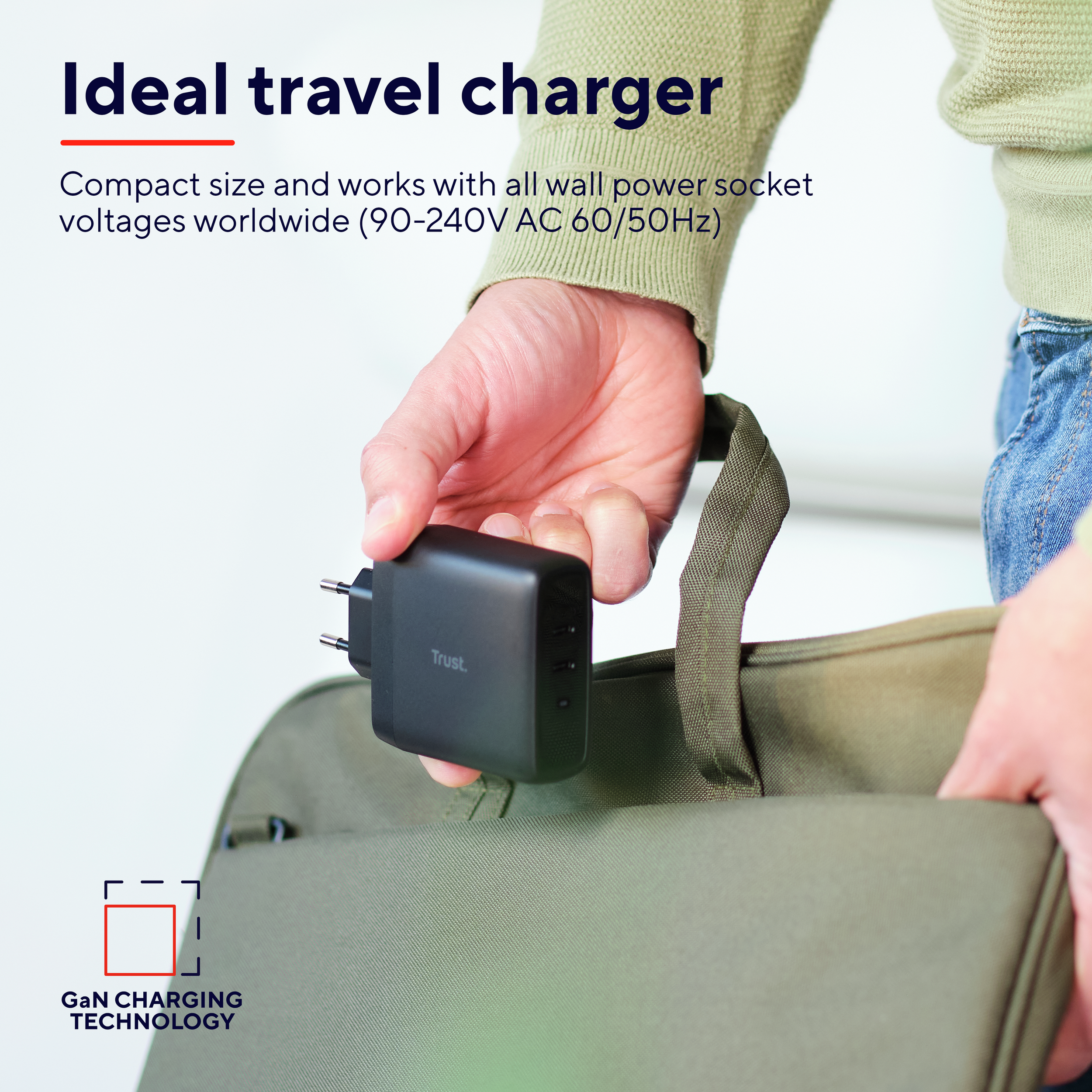 De hand van een persoon die een zwarte oplader vasthoudt met een tas op de achtergrond.