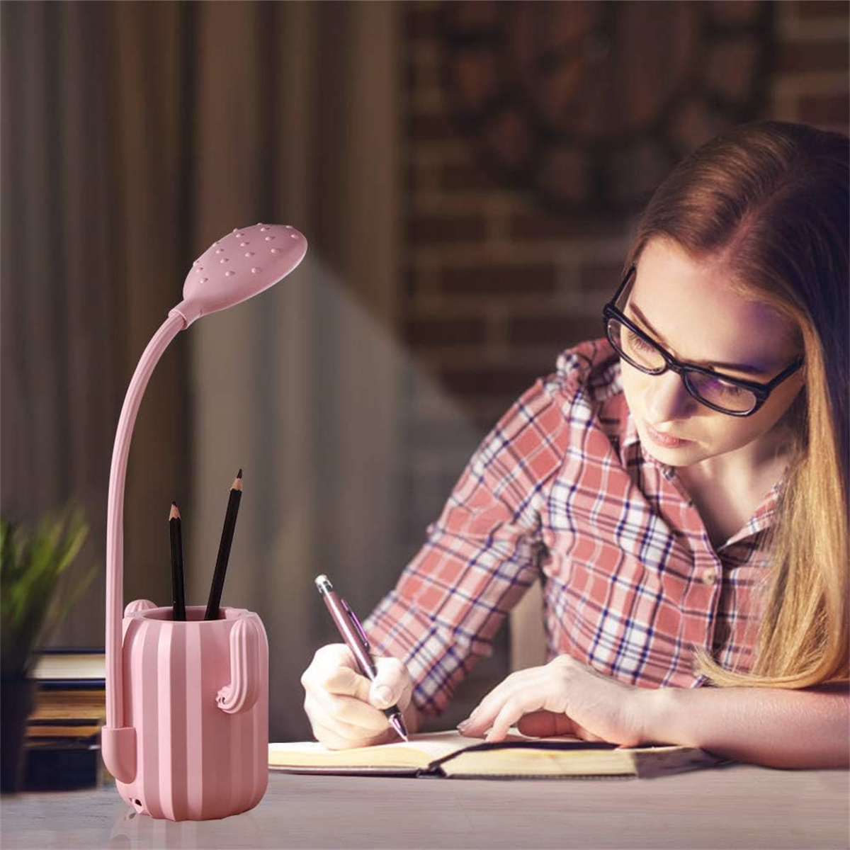 Eine Frau mit Brille schreibt in einem Notizbuch an einem Schreibtisch. Eine rosa Schreibtischlampe mit Stifthalter steht auf dem Tisch.