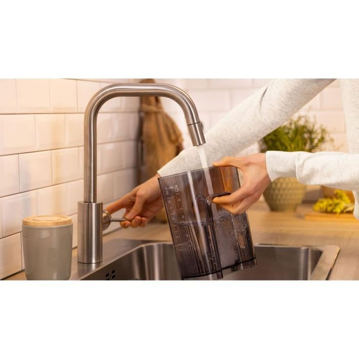 Osoba napełniająca przezroczysty pojemnik wodą z kranu. W tle zlewozmywak.