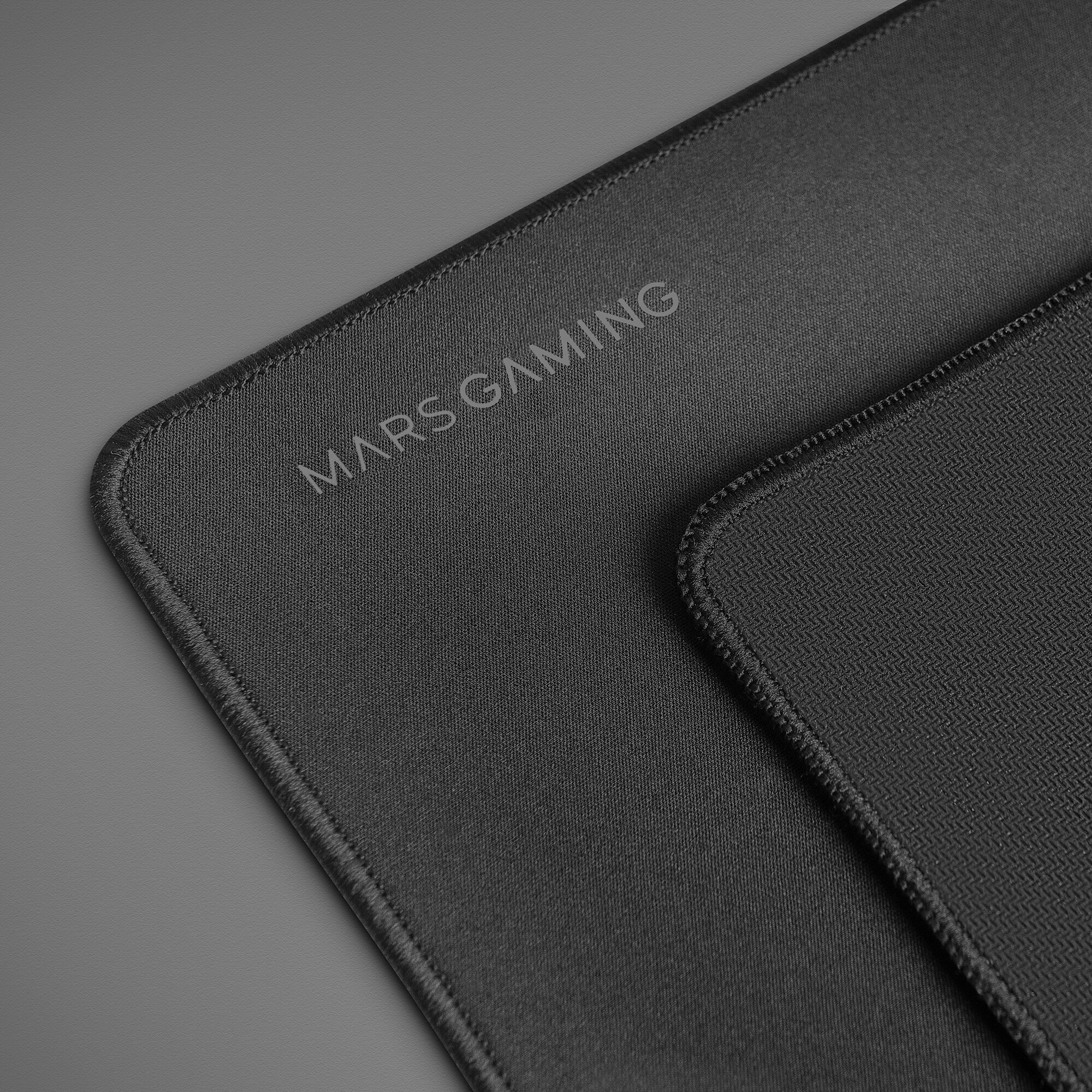 Primer plano de una alfombrilla de ratón gaming negra con MARS GAMING. Una alfombrilla más pequeña en el fondo.
