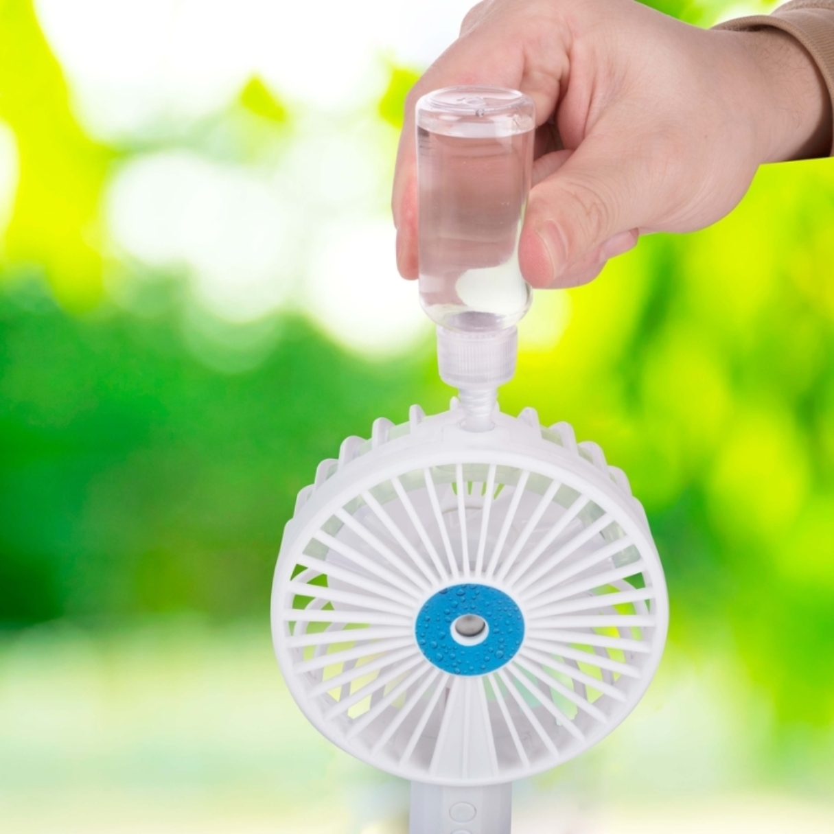Une main versant du liquide dans un ventilateur portable blanc avec un centre bleu.
