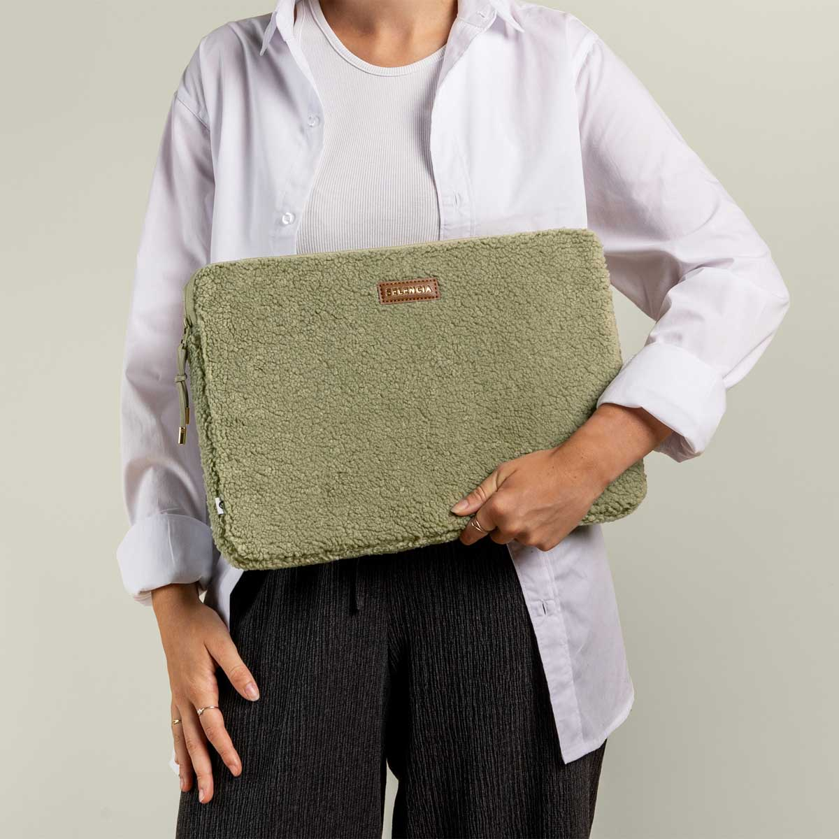 A person holding a green laptop sleeve with a label 'SELENCIA.' The person is in a white shirt and black pants.