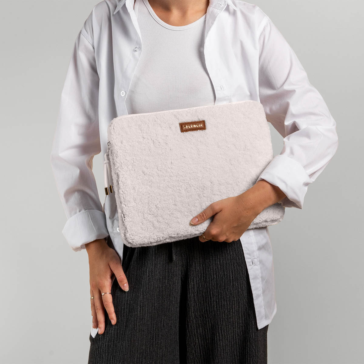 A woman holds a white fuzzy laptop case. She wears a white shirt and black pants.