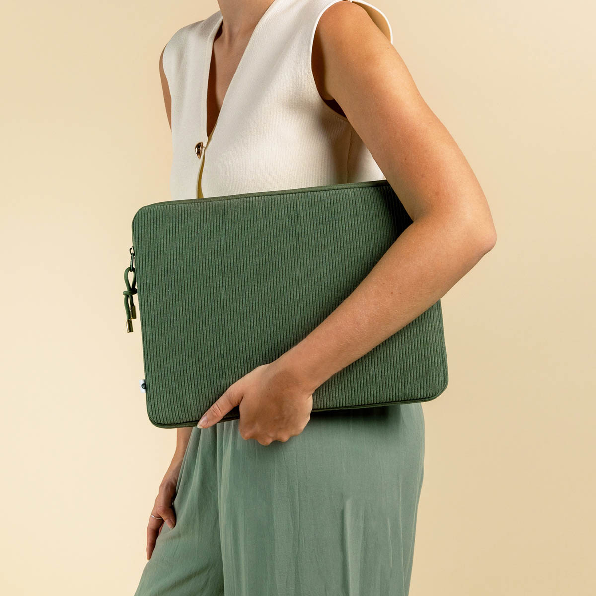A woman holding a green corduroy laptop sleeve. She is wearing a white top and green pants. The background is beige.