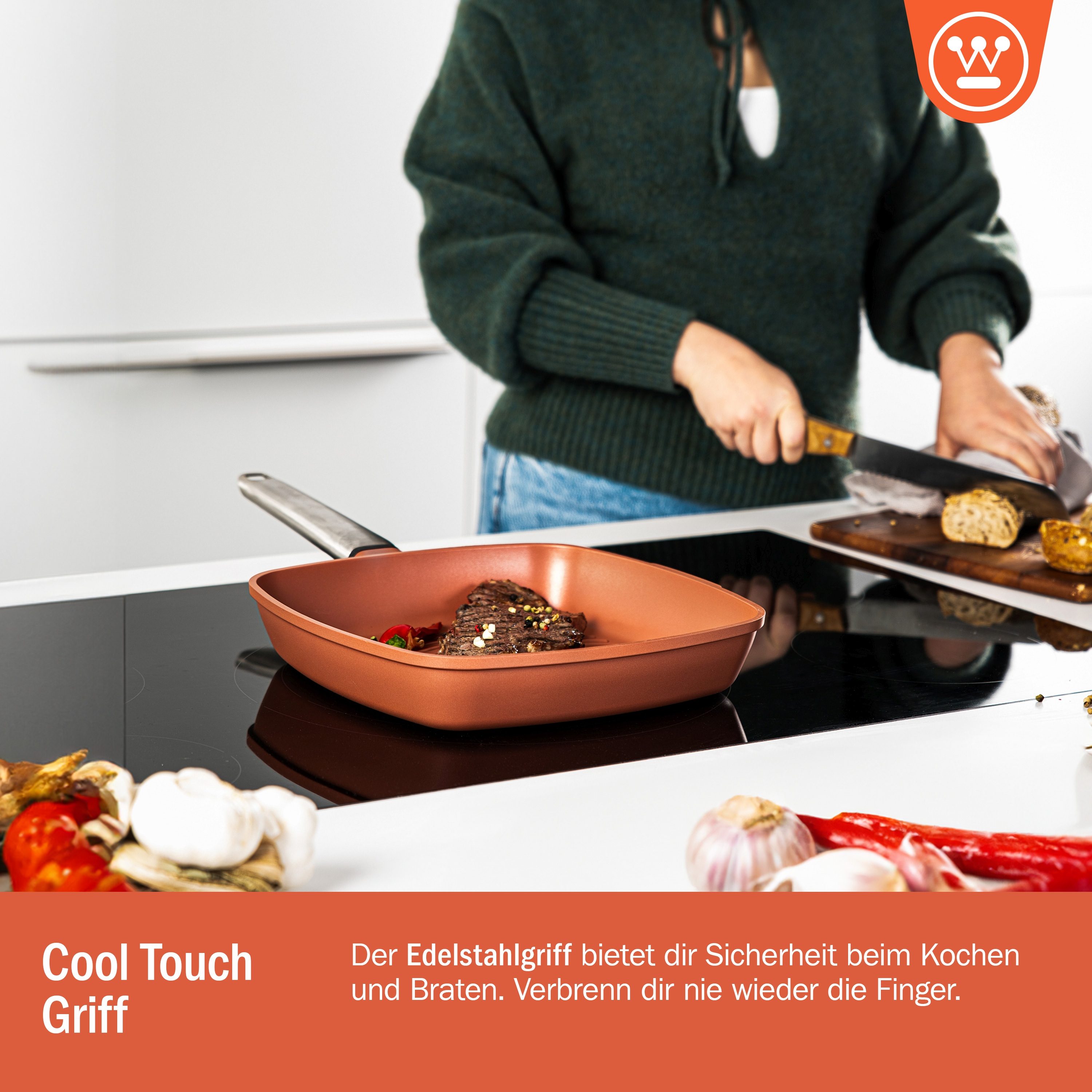 Eine Kupferpfanne mit Essen auf einem schwarzen Herd, eine Person schneidet Brot. Knoblauch und rote Paprika sind im Vordergrund.